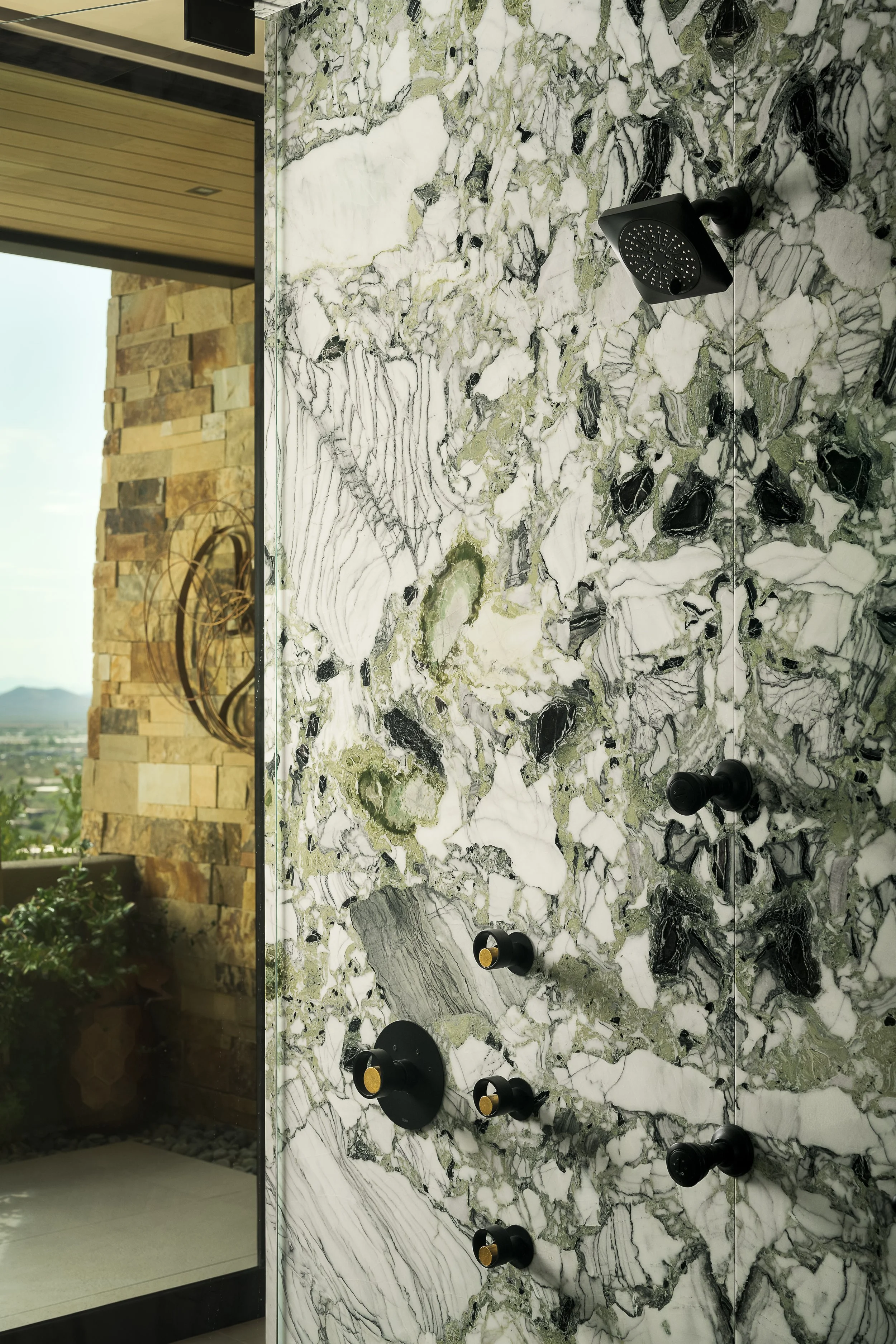 Close-up of a marble shower wall with black shower fixtures, located next to a glass window that shows a stone wall and outdoor landscape.