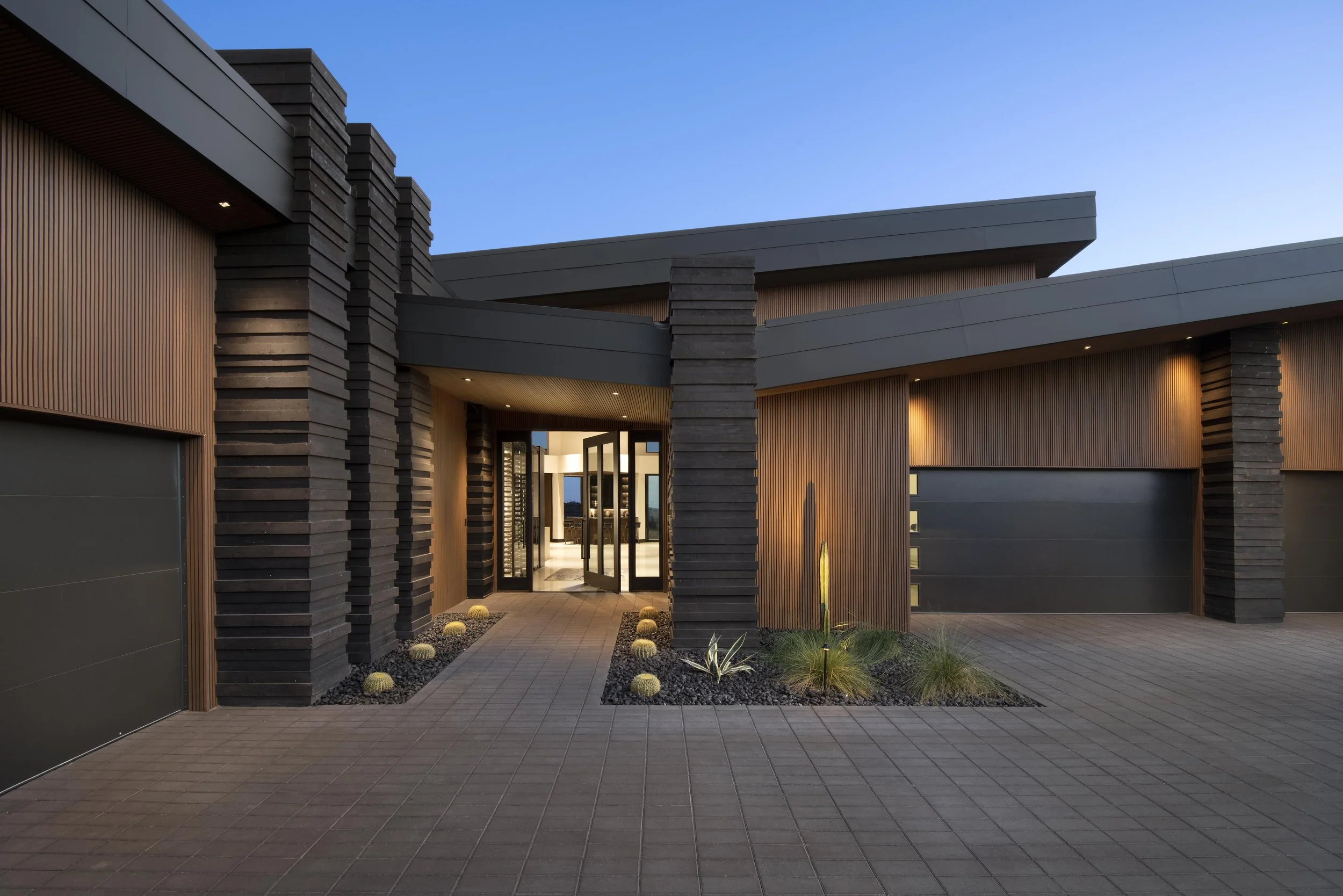 Modern house with wooden and dark gray exterior, illuminated entrance, landscaped pathway with desert plants, and a clear blue sky at dusk.