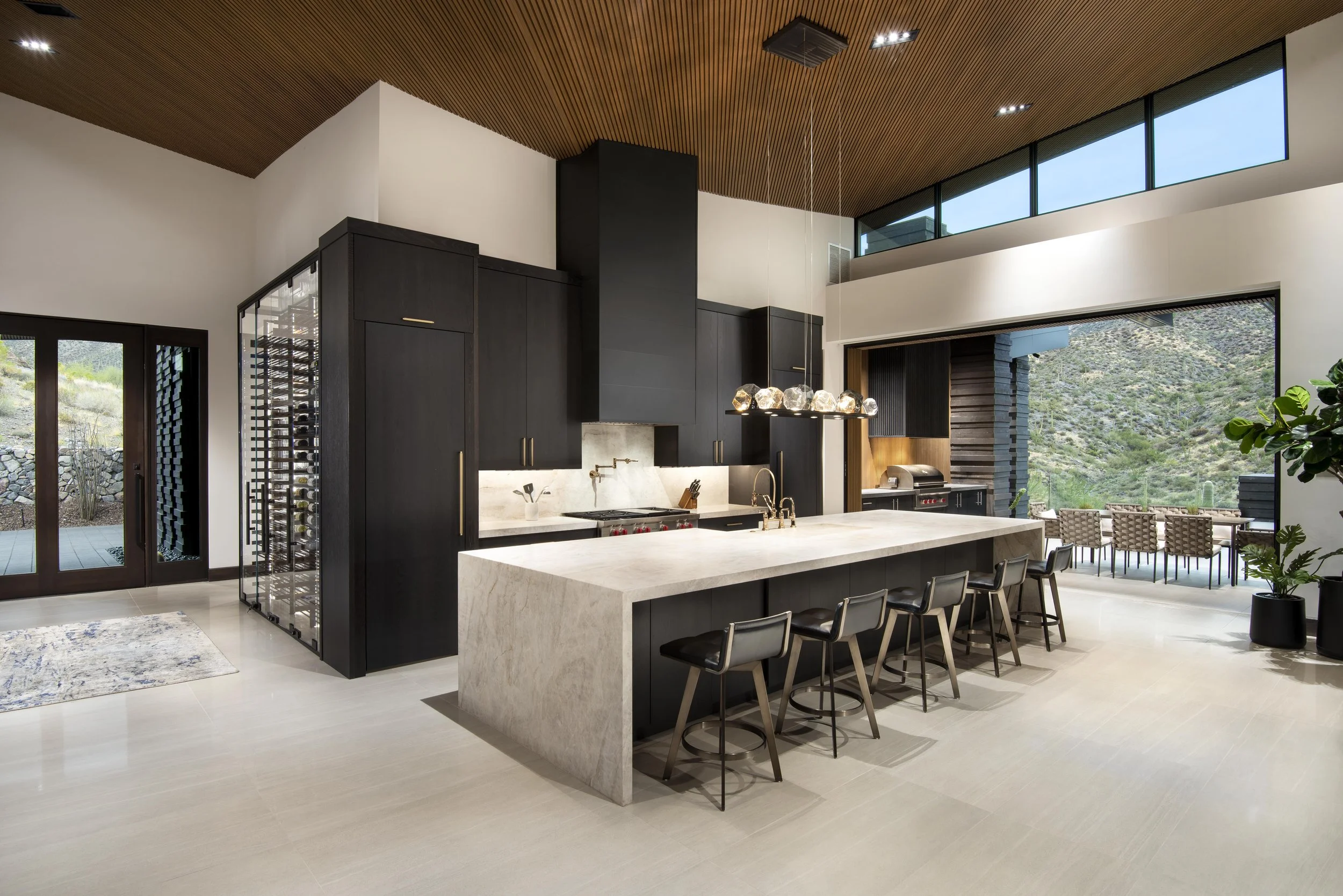 Modern open-concept kitchen with a central island, black oak cabinets, a Taj Mahal countertop, a built-in Wolf Subzero oven, a large built in wine wall, and large windows showing an outdoor landscape.