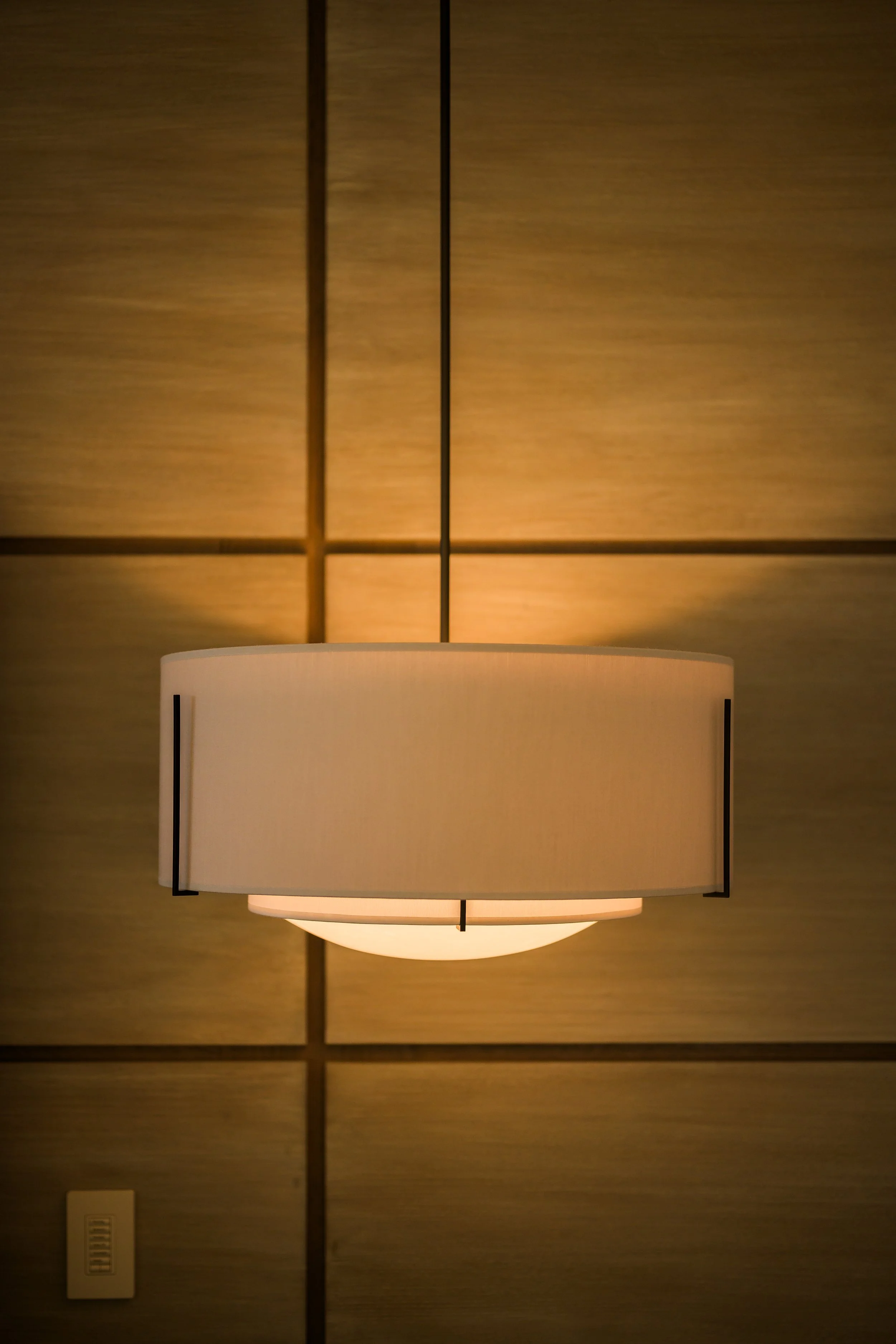 A modern wall-mounted lamp with a cylindrical white shade, illuminated, affixed to a beige wall with wood paneling.