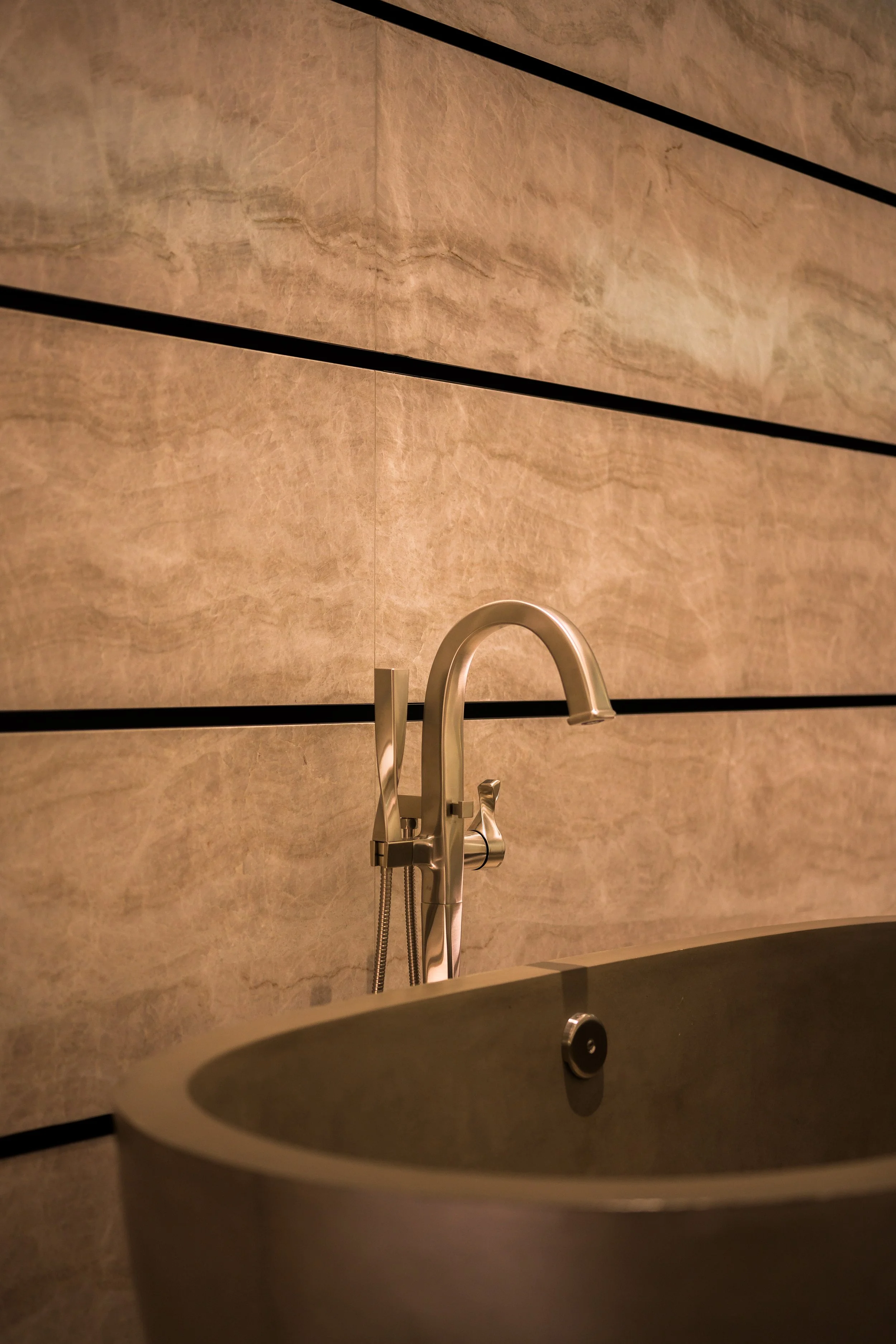 Close-up of a modern bathroom faucet and bathtub against a beige marble wall.
