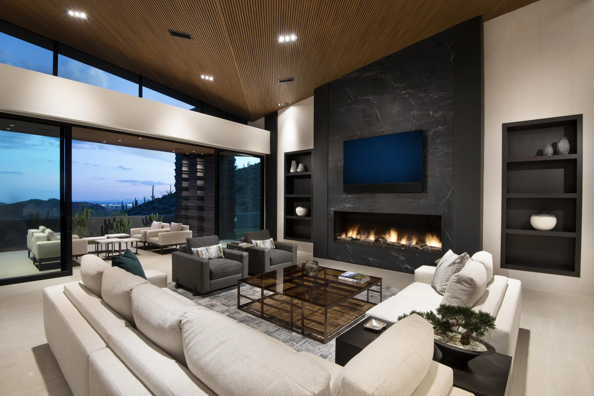 Modern living room with large beige sectional sofa, dark gray armchairs, glass coffee table, fireplace with dark marble surround, black shelves with decorative items, large window with balcony overlooking a desert landscape, wood-paneled ceiling, and a prominent flat-screen TV.
