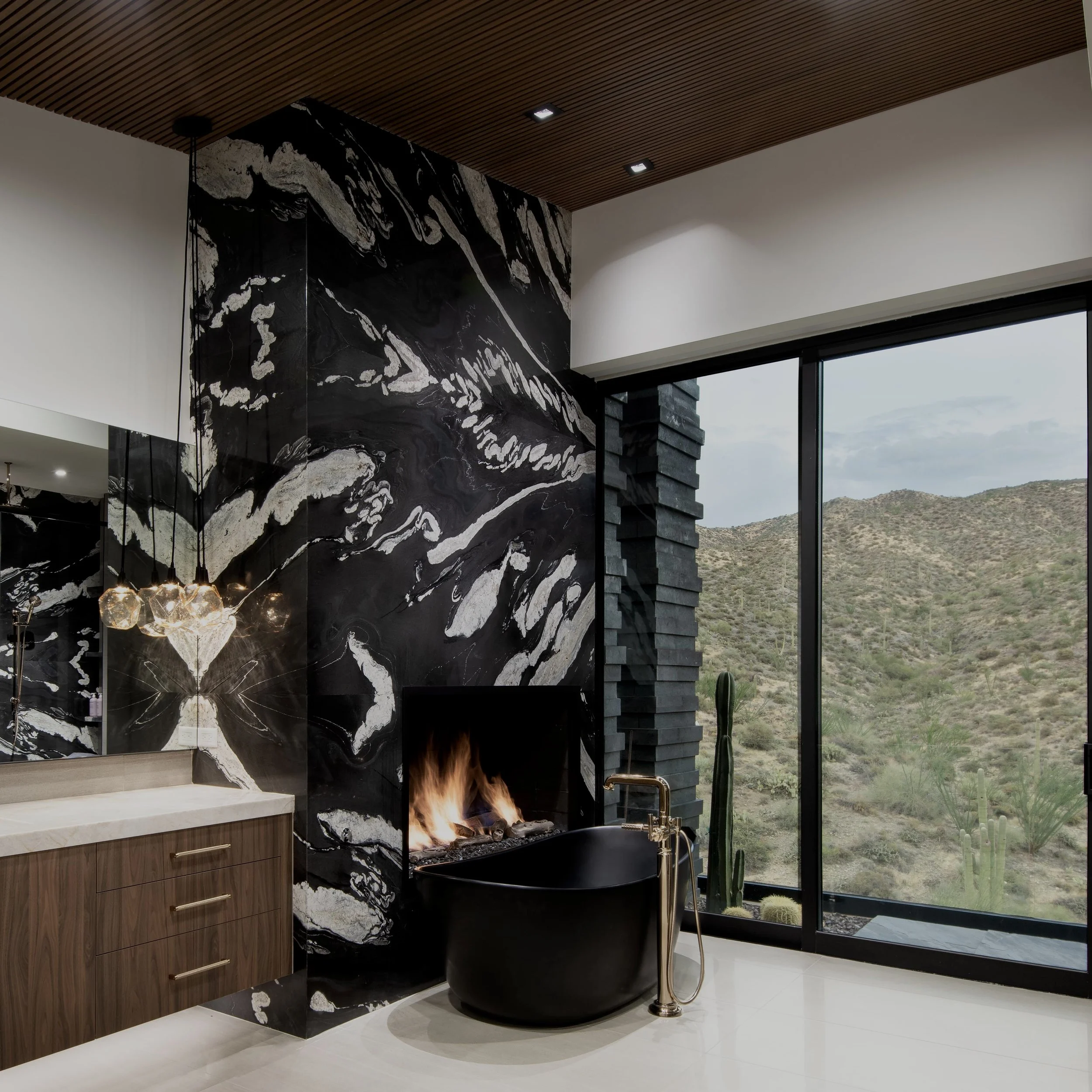 Modern interior master bath with black marble fireplace,  free standing matte black bathtub, walnut cabinetry, large floor-to-ceiling windows showing desert landscape.