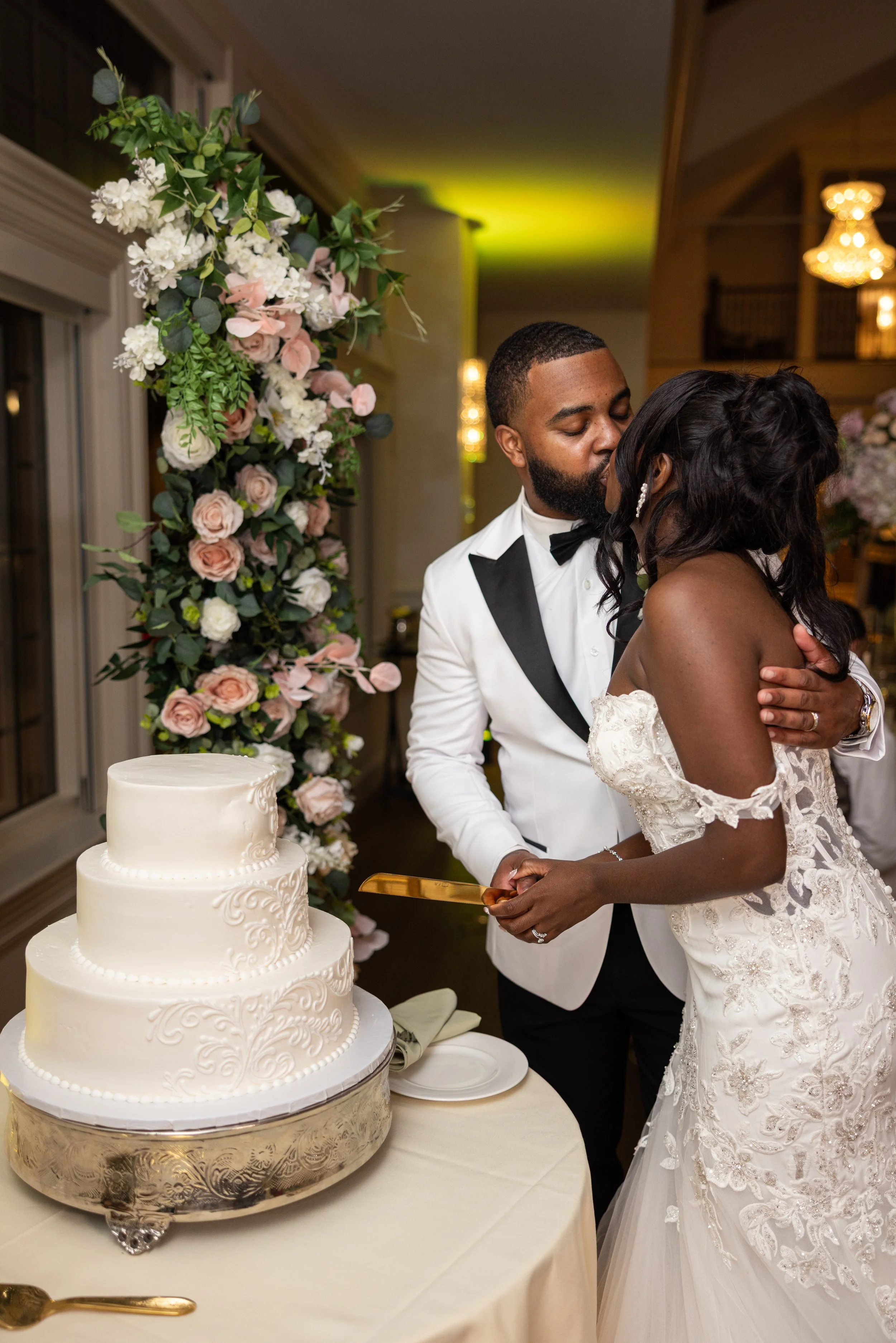 A couple in wedding attire sharing a kiss during their wedding reception, with a three-tiered white wedding cake on a table in front of them, decorated with a floral arch in the background.