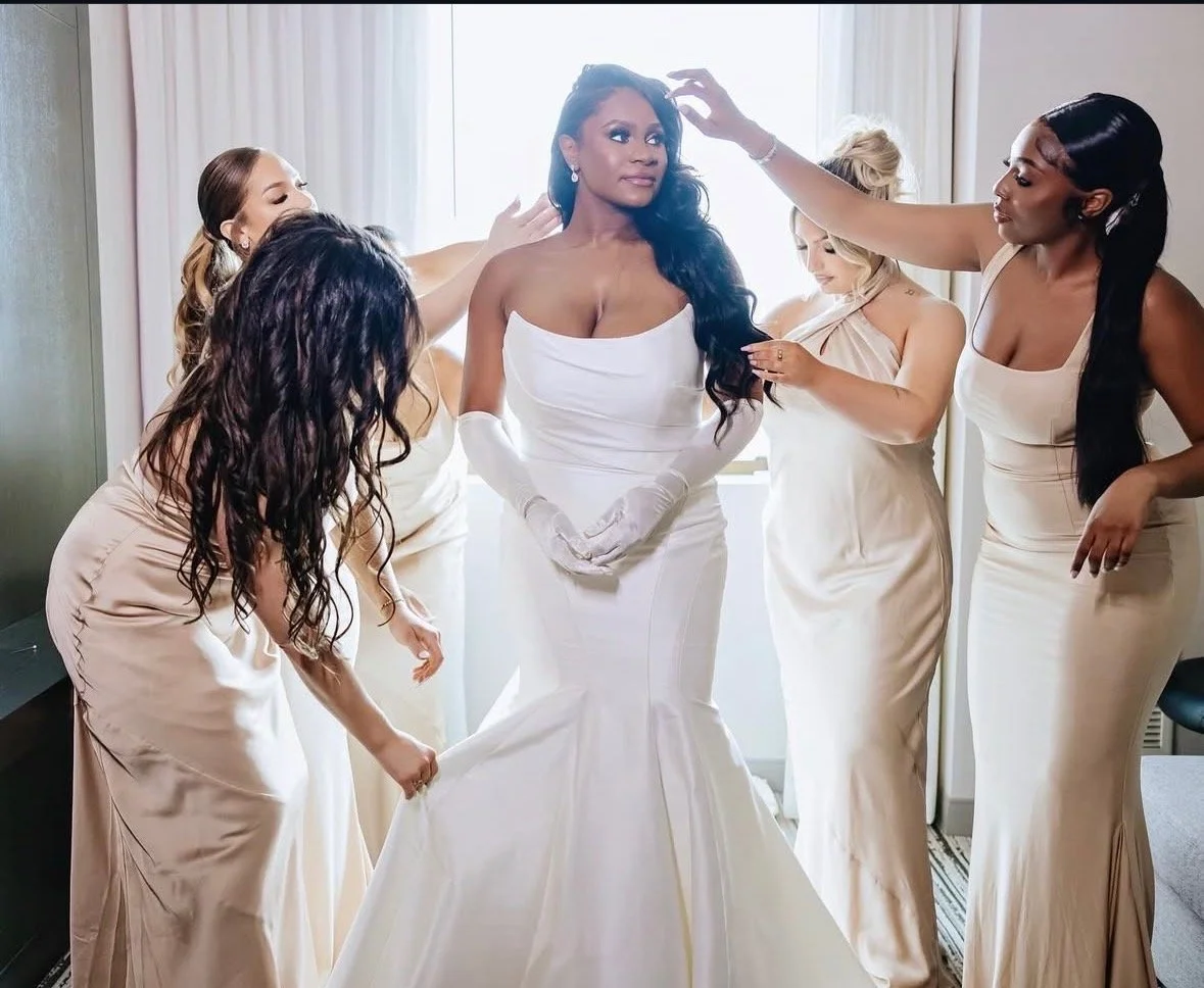 A bride in a white wedding gown with long gloves being helped by five bridesmaids in matching champagne-colored dresses as they prepare for her wedding.