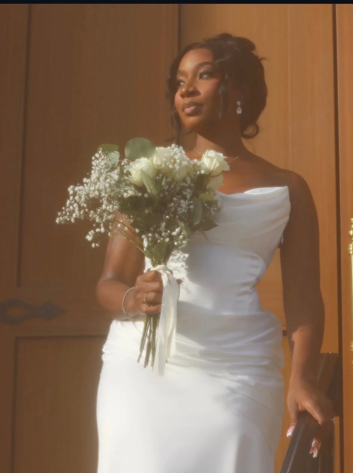 A bride in a white wedding dress holding a bouquet of white roses and baby's breath, standing in front of a wooden door.