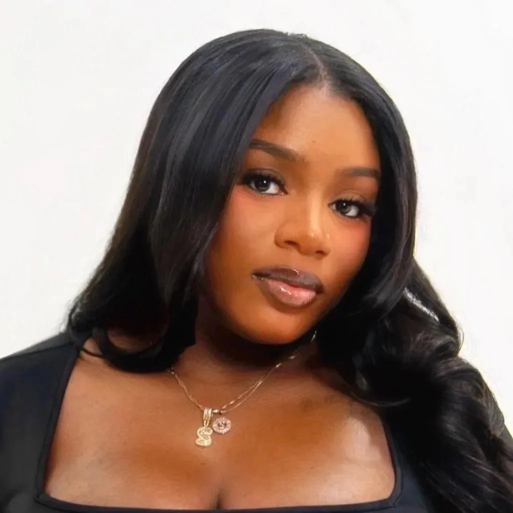 Portrait of a young woman with long black hair, wearing a black top and a gold necklace, posing against a plain white background.