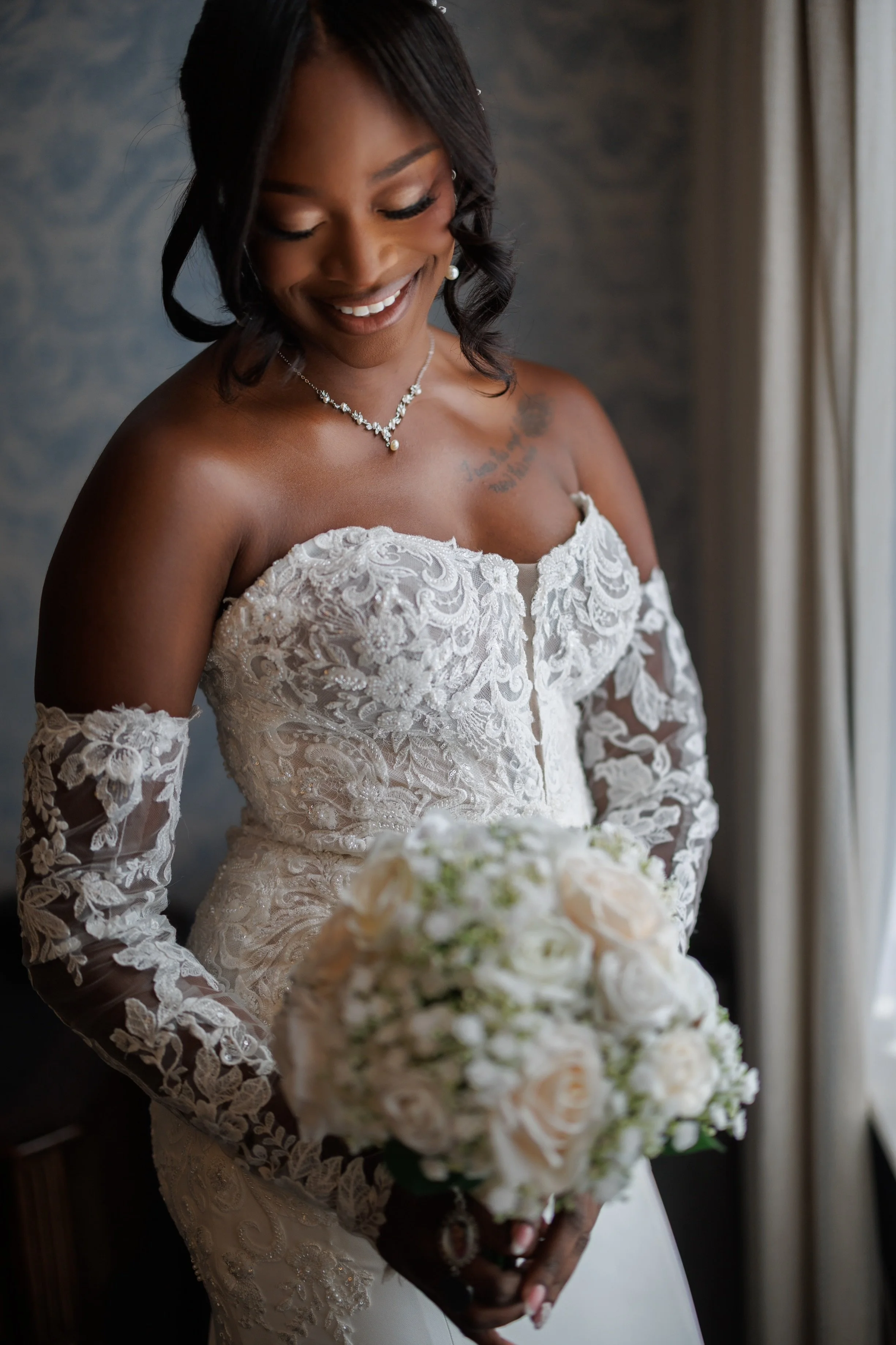 A bride in a lace wedding dress holding a bouquet of white roses and peonies, smiling and looking down, standing near a window with curtains.