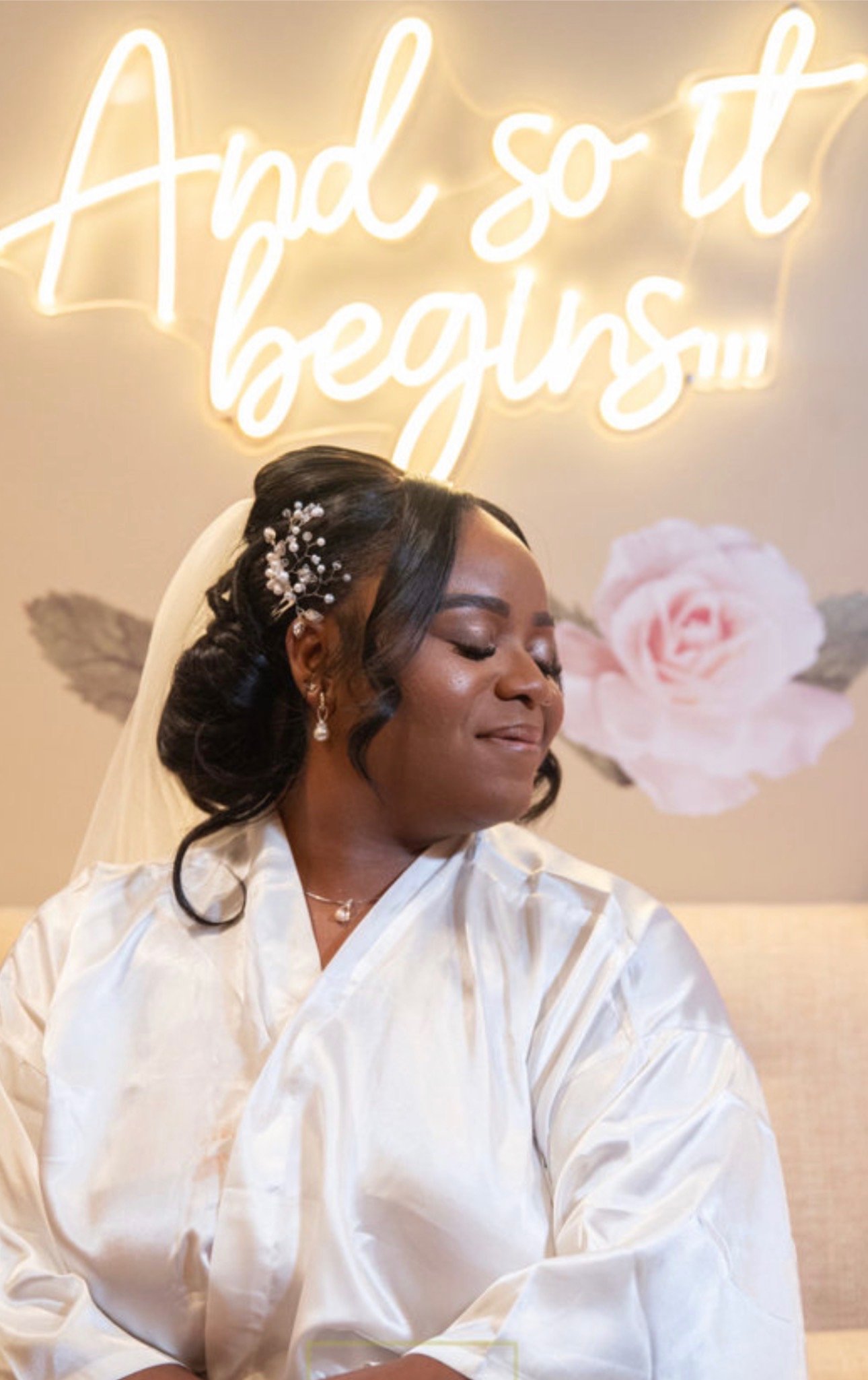 A bride dressed in a white satin robe with a veil, her hair styled in an elegant updo with pearl hair accessories. She is smiling softly with her eyes closed, standing in front of a wall with a neon sign that reads 'And so it begins...' and a pink floral decoration.