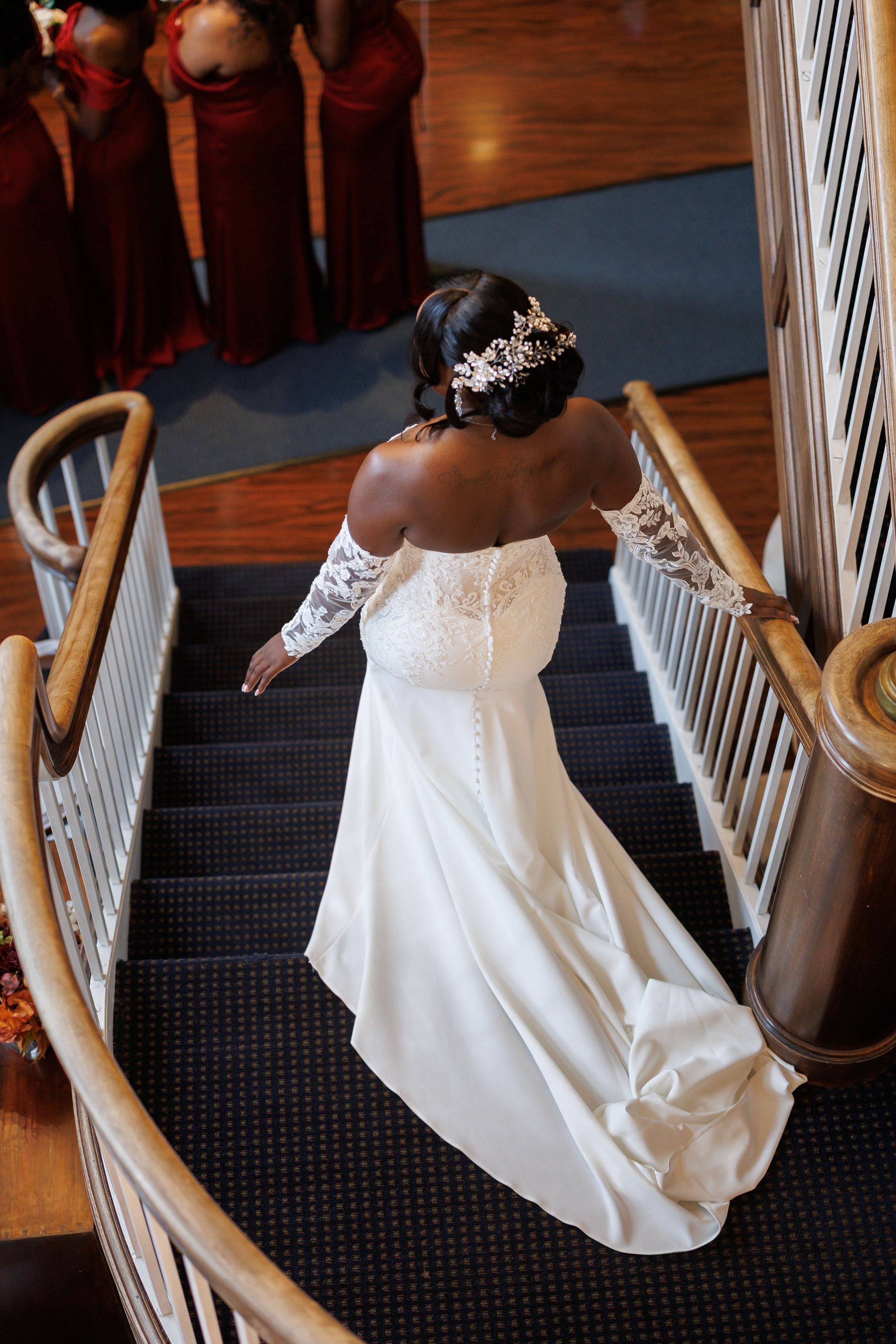 A bride ascending a staircase wearing a strapless wedding gown with lace sleeves and a floral headpiece, with bridesmaids in burgundy dresses standing at the top.