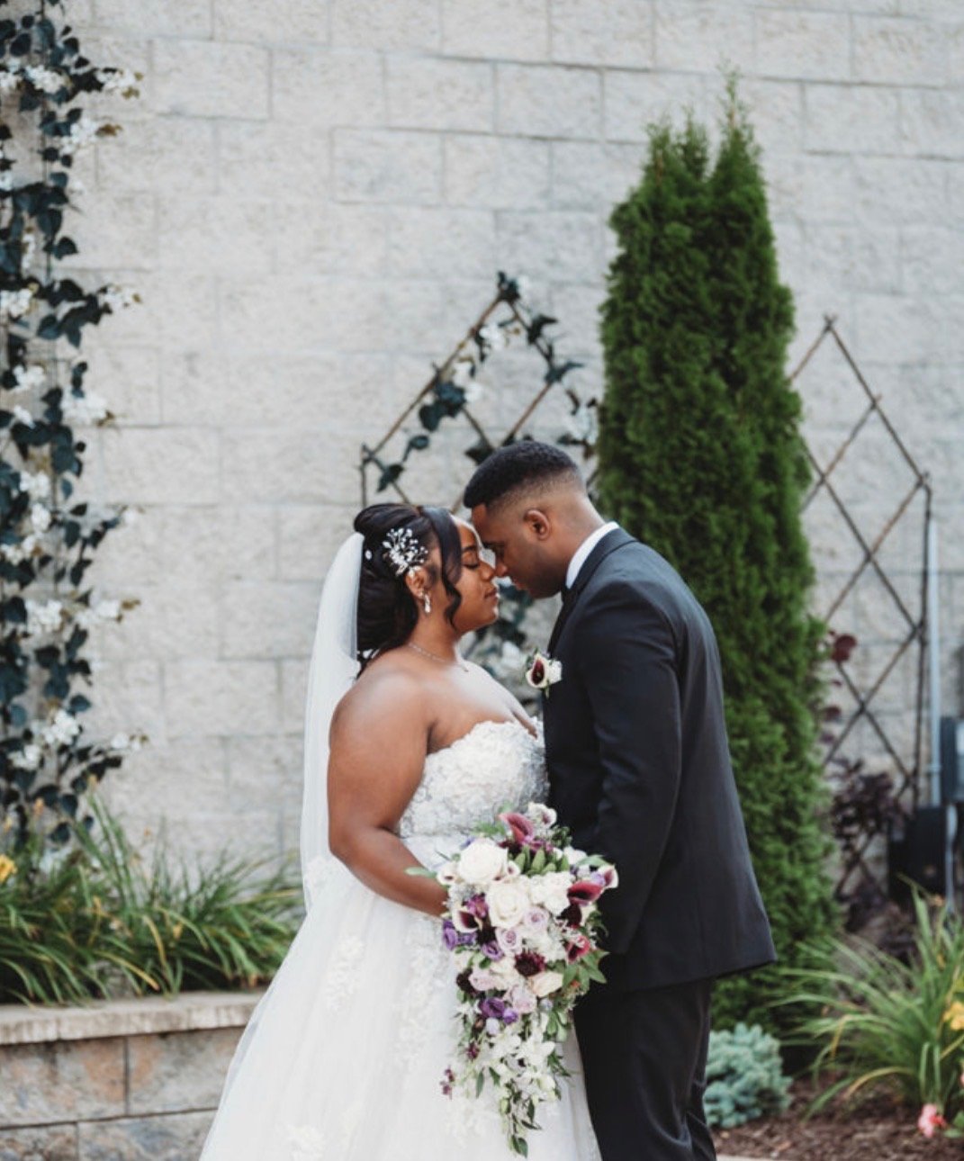 A bride and groom stand close together with foreheads touching, outdoors in front of a wall and greenery, the bride holding a cascading bouquet of flowers.