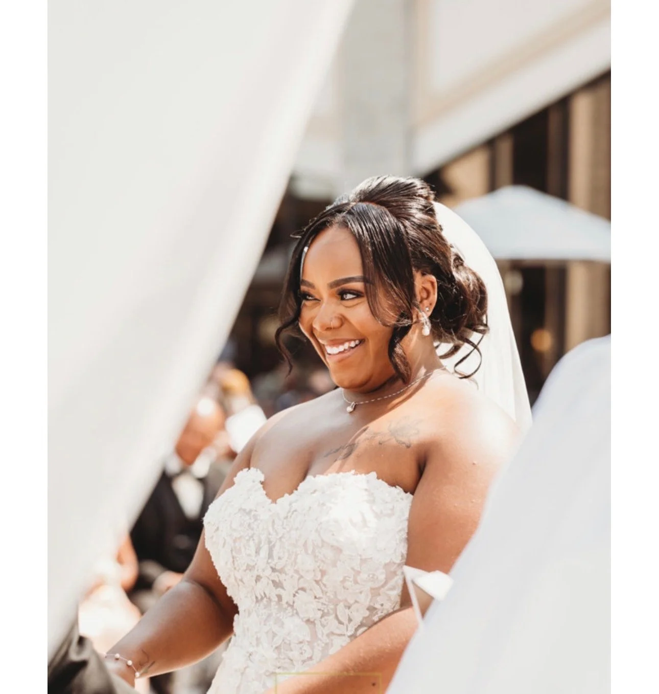 Bride smiling during her wedding ceremony, wearing a strapless white wedding dress with lace detailing, veil, and jewelry, outdoors with guests and umbrellas in the background.