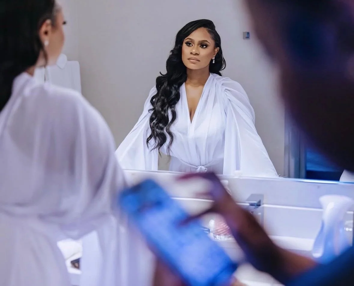 A woman with long, wavy black hair, dressed in a white outfit, is looking at a reflection in a mirror while two other women, also in white outfits, are present in the foreground, one of whom is using a mobile phone.