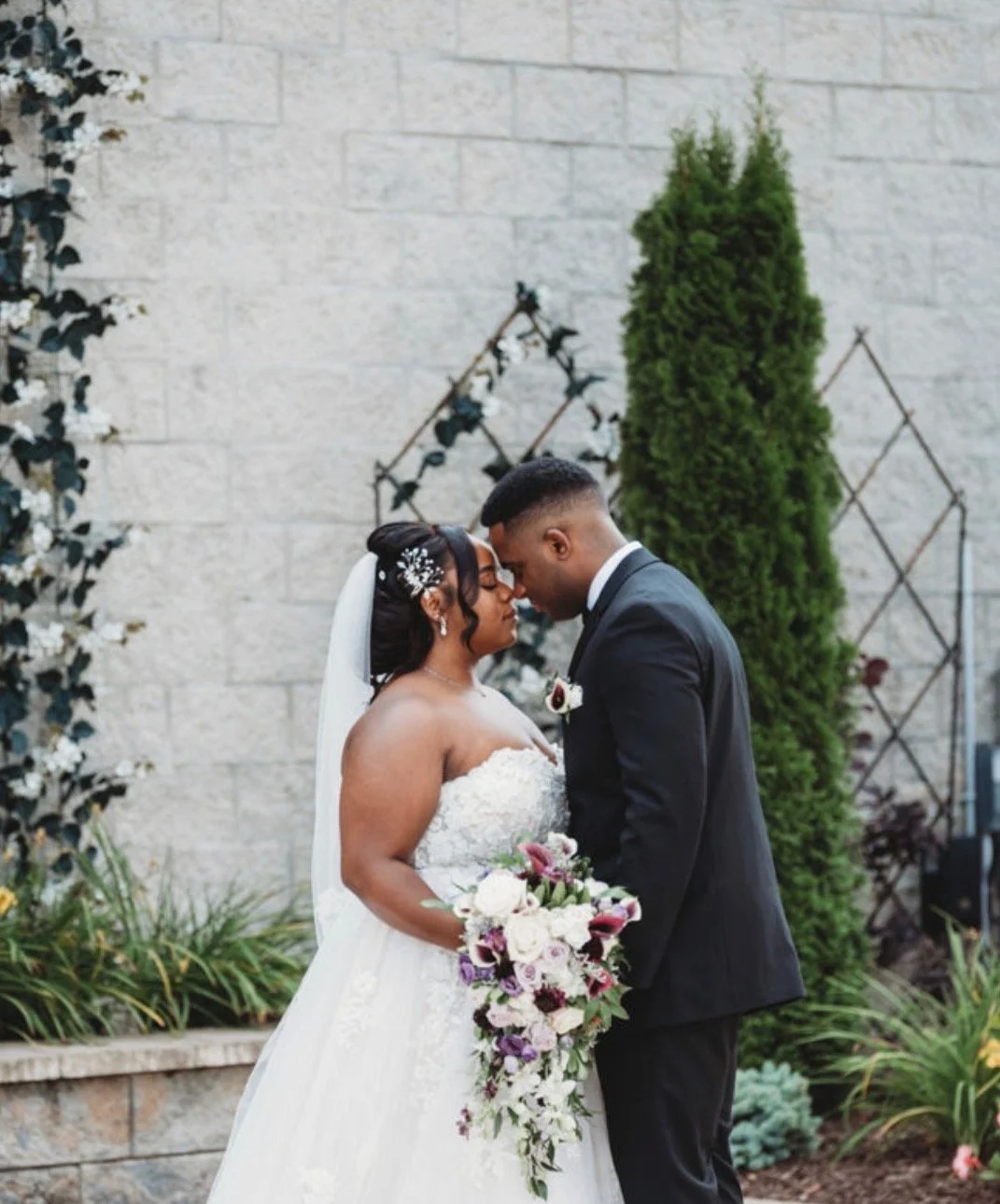 Bride and groom standing close together, forehead touching, outdoors with greenery and a wall.