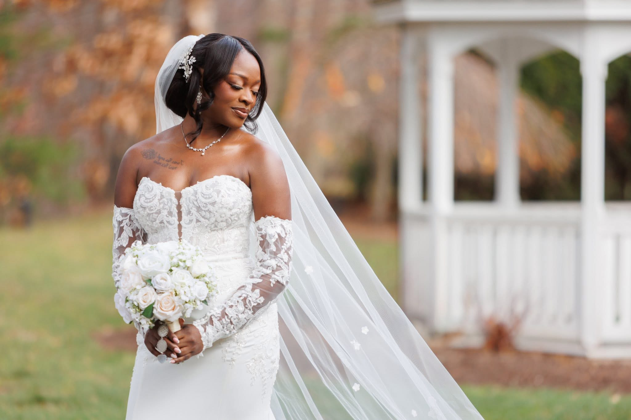 Bride in a white wedding dress holding a bouquet of white roses, standing outdoors with a white gazebo in the background.