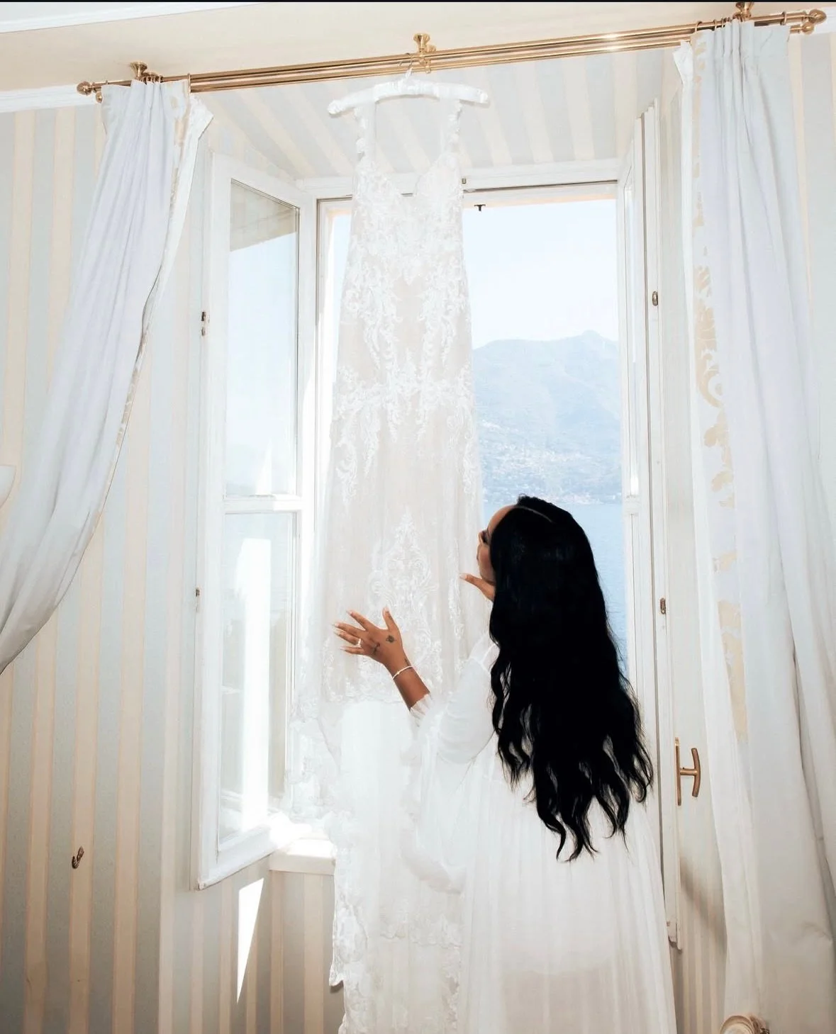 A woman with long black hair standing near an open window, touching a hanging white lace wedding dress, with a mountain and water view outside.