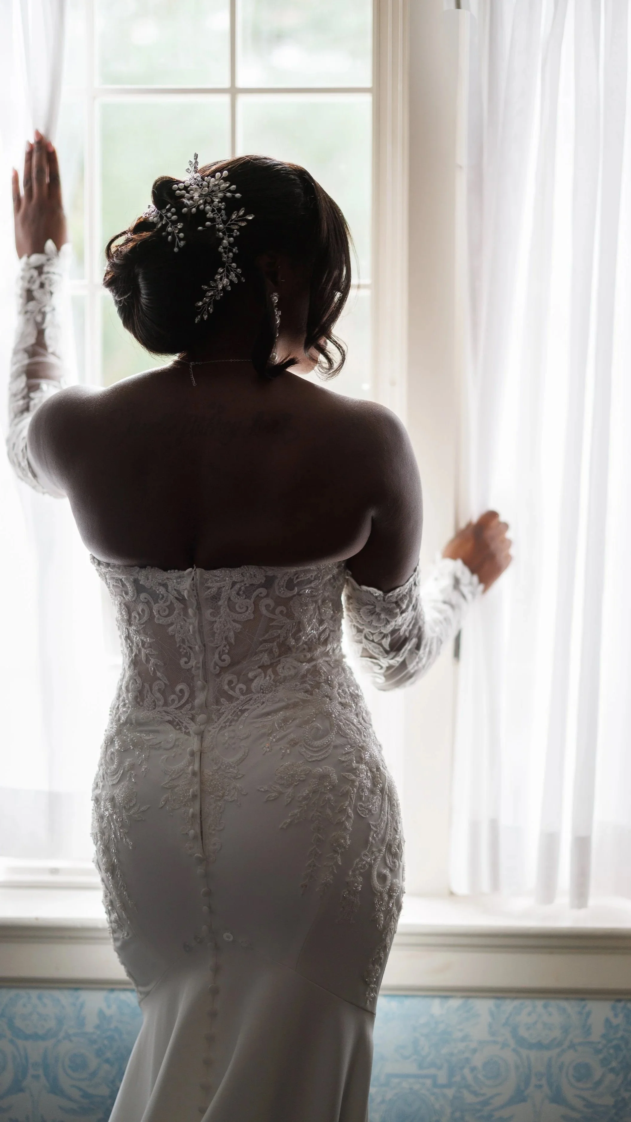 A bride in an off-the-shoulder lace wedding gown standing by a window, looking outside with her hand on the curtains.
