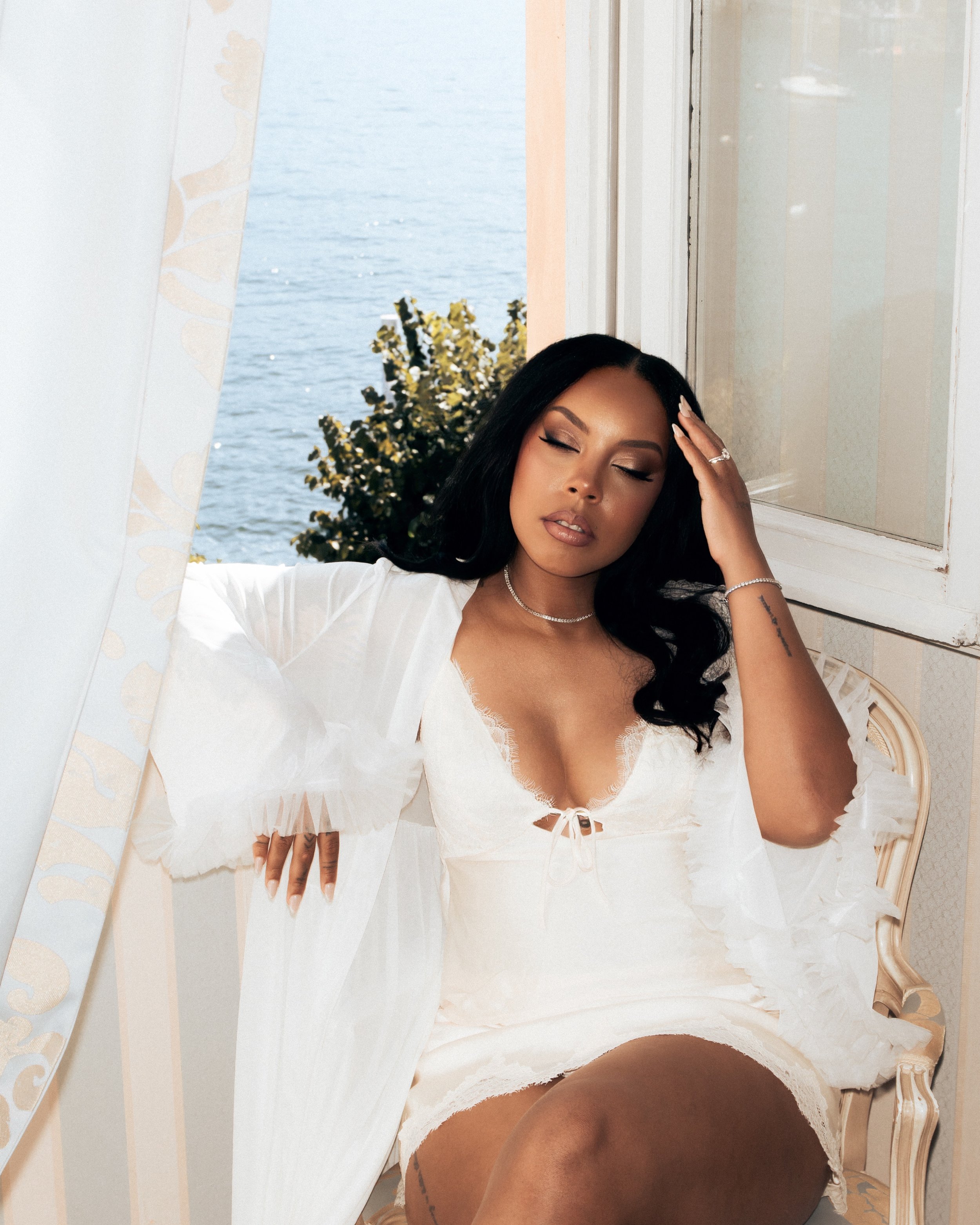 A woman with long black hair, sitting on a vintage chair near a window, wearing a white dress and jewelry, with a body of water visible outside.
