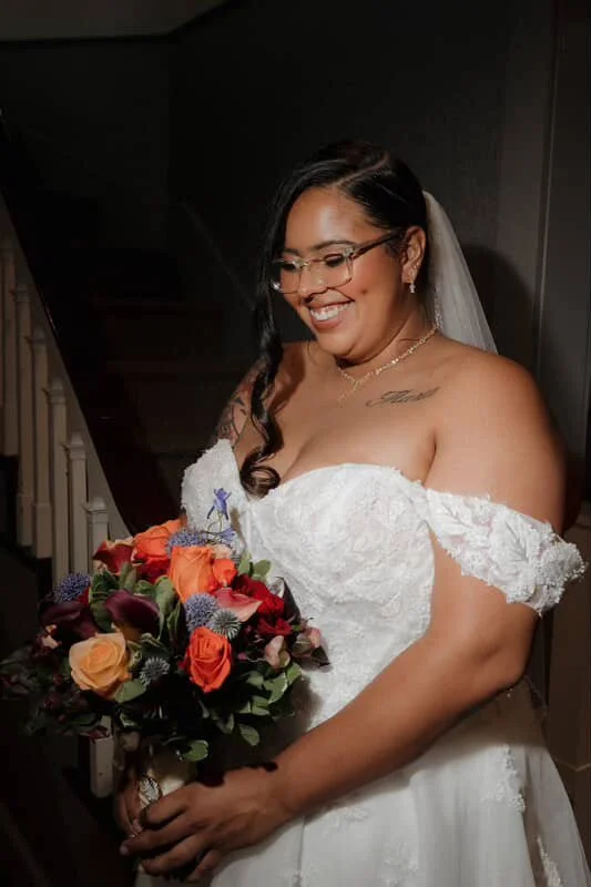 A bride in a white wedding dress holding a colorful bouquet of flowers, smiling and looking down.