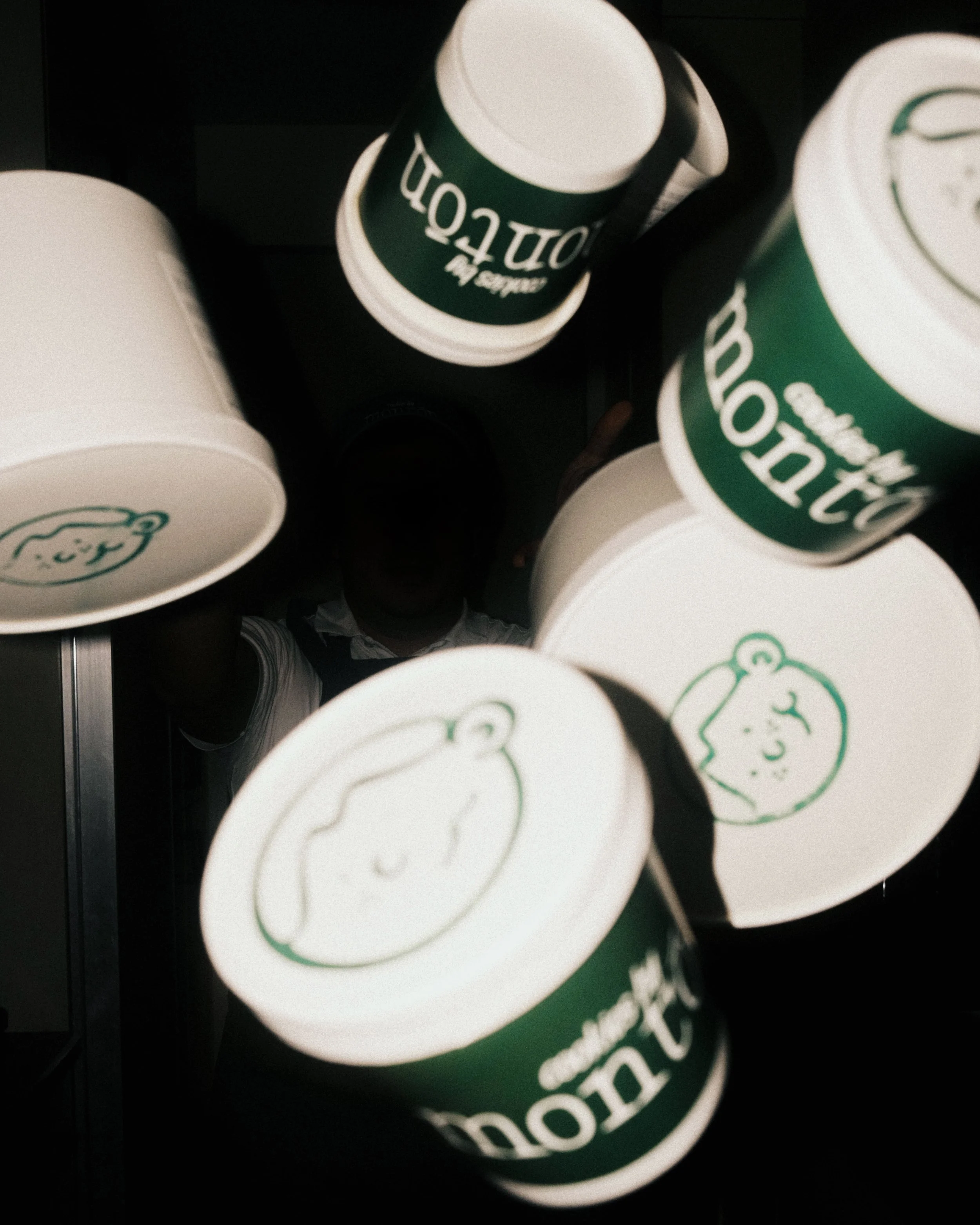 Close-up view of several disposable cups with a green and white logo featuring a cute smiling face, with an individual in the background holding a coffee cup.