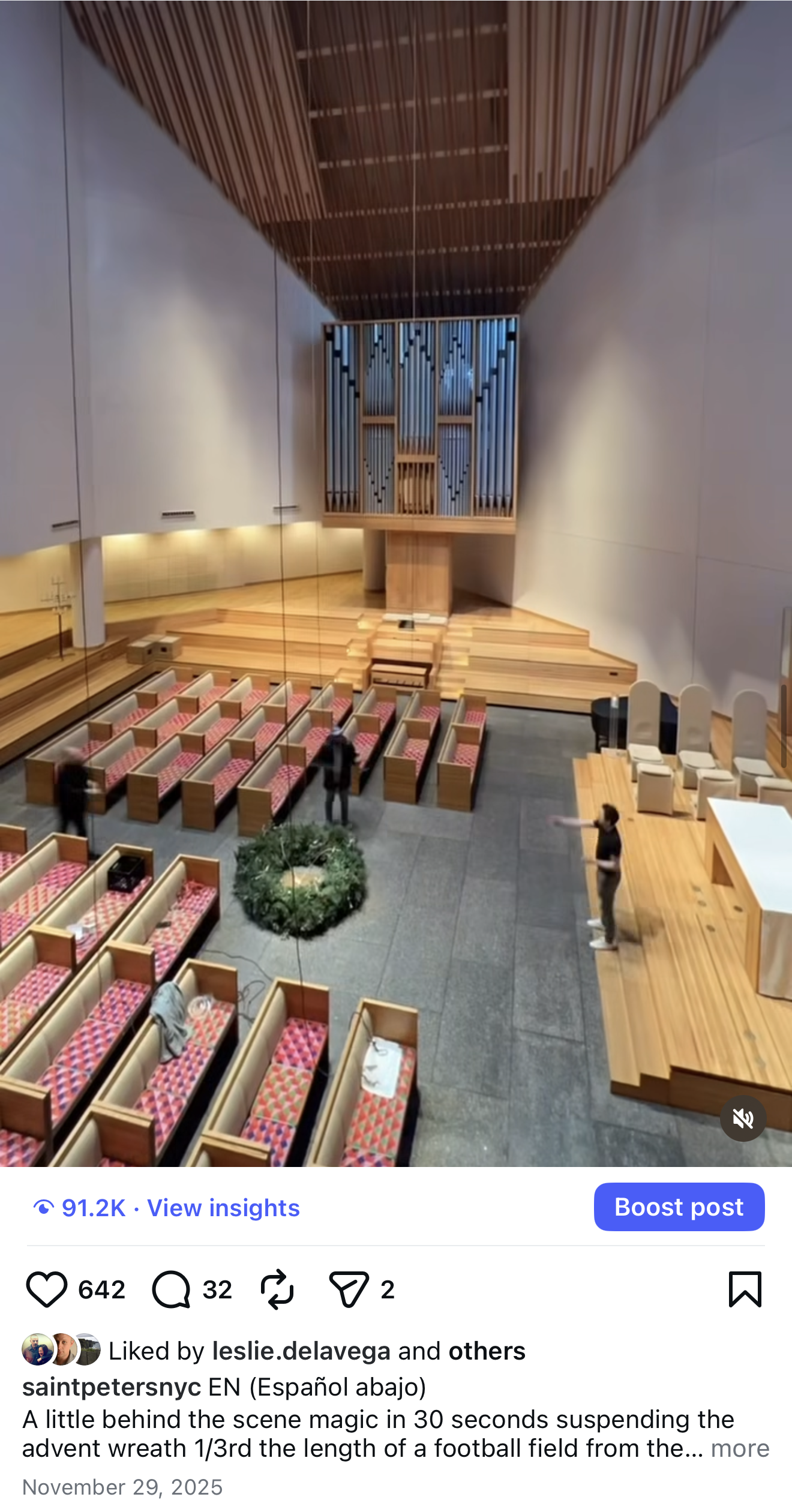 Interior of a church or chapel with wooden pews arranged in rows, a stage area to the right with white chairs, and a large pipe organ at the back. The ceiling is wooden with horizontal slats, and there are three people inside the space.