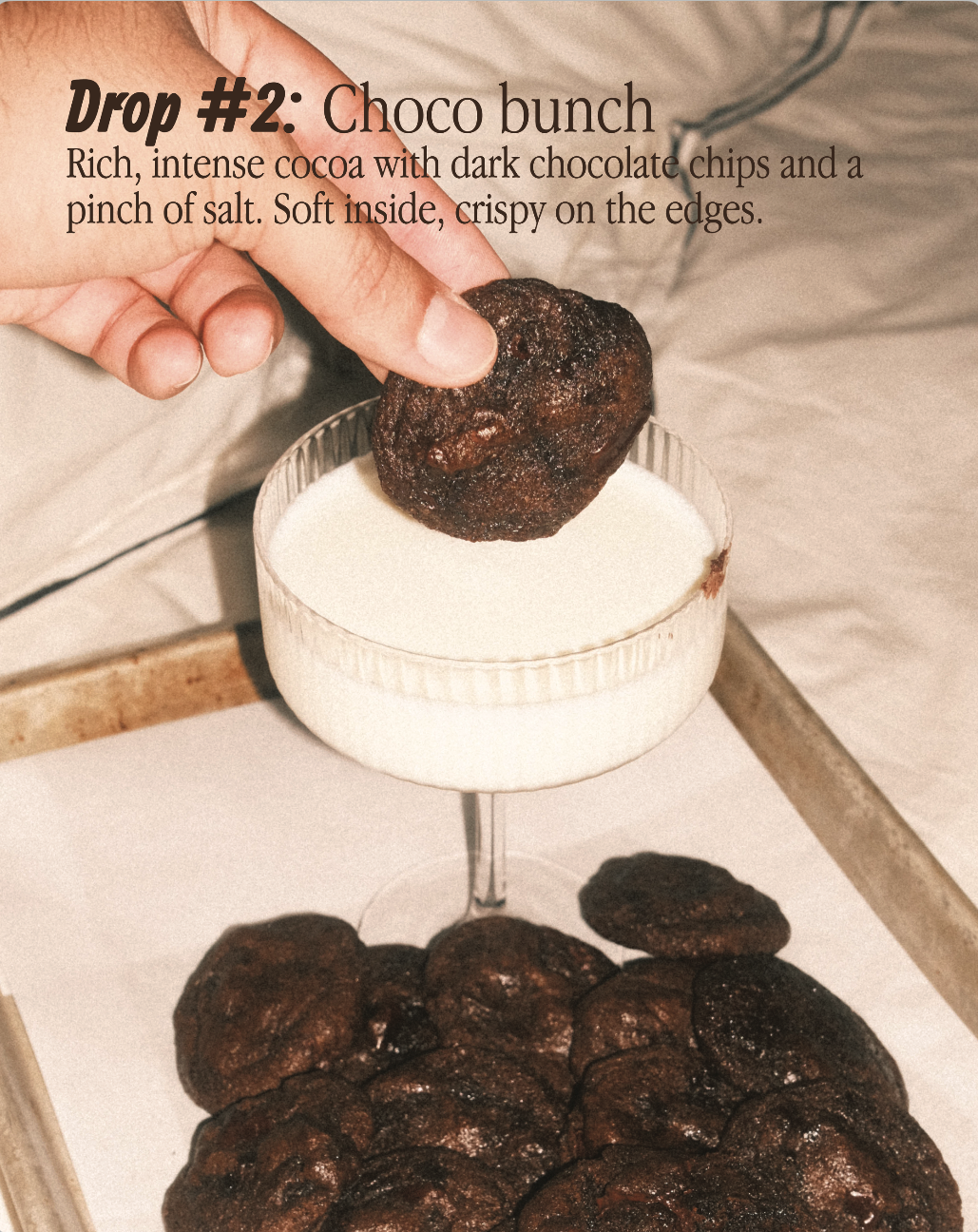 Close-up of a hand holding a chocolate cookie over a white bowl filled with milk. Several more dark, crispy chocolate cookies are on a tray below.