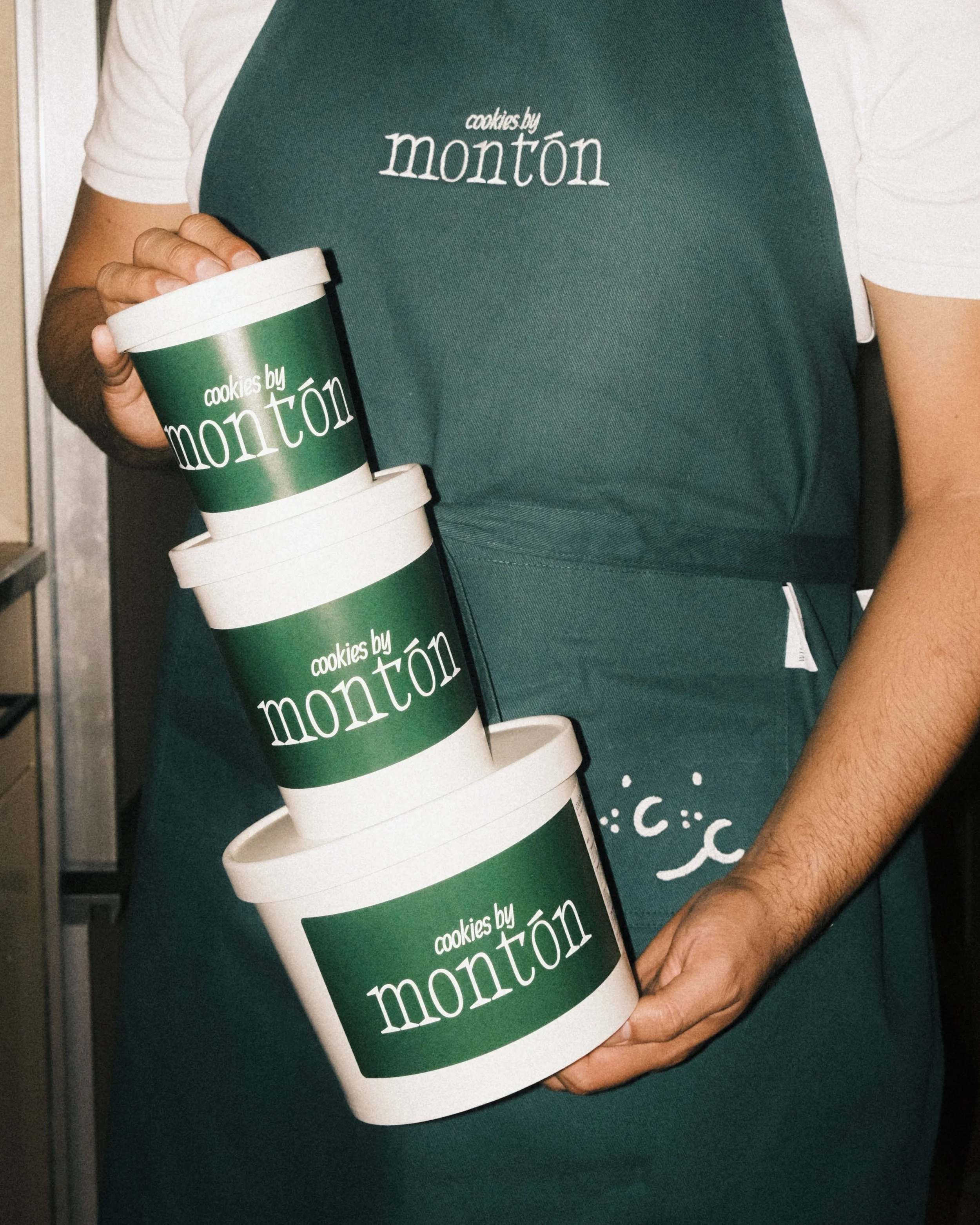 Person holding three stacked containers of cookies from cookies by Monton, wearing a teal apron with the same logo.