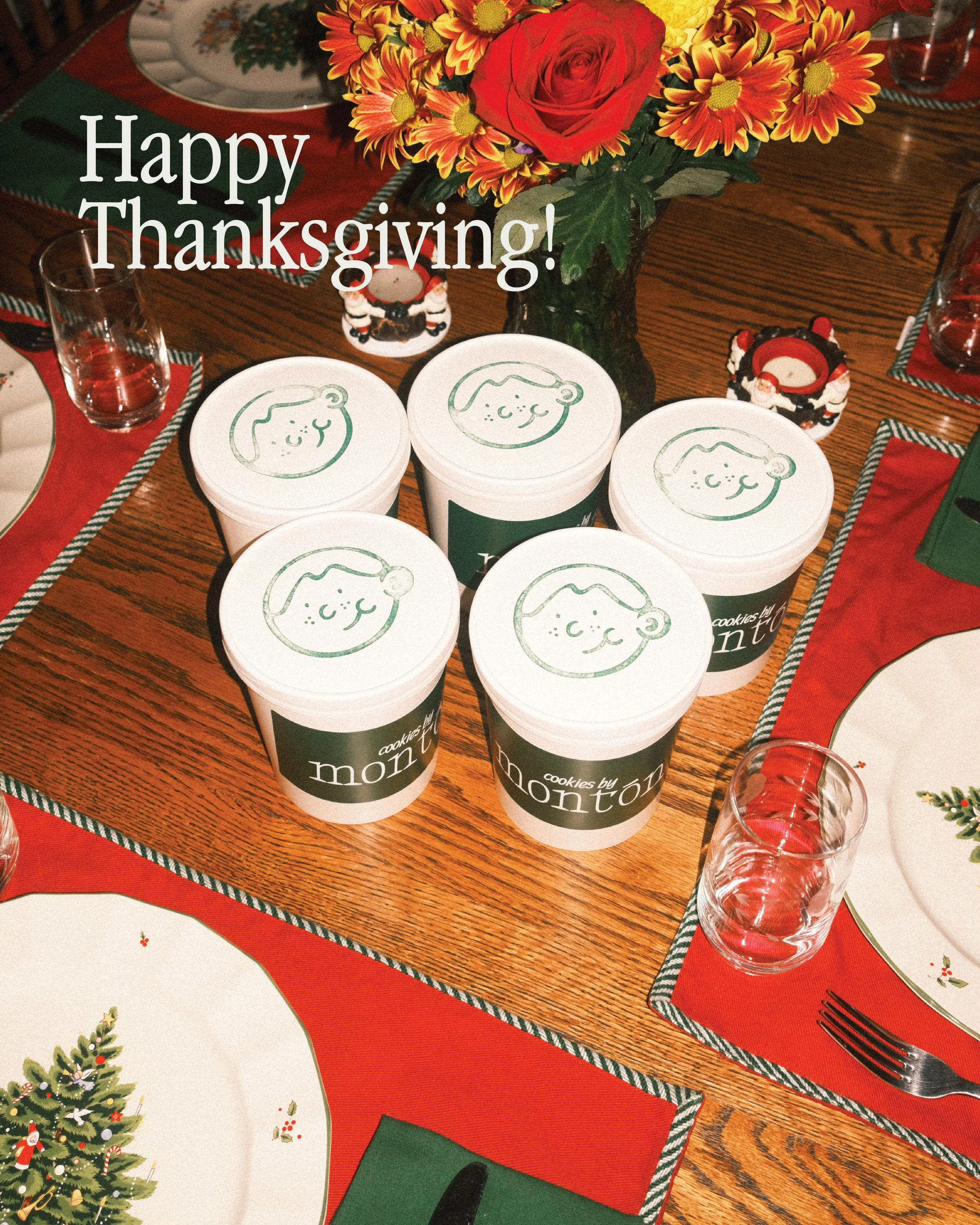 A holiday dinner table with a center vase of red, yellow, and orange flowers, surrounded by festive tableware including Santa-themed cookie jars labeled "cookies by monti," drinking glasses, plates with Christmas tree designs, napkins with holly decorations, and a "Happy Thanksgiving!" message overlay.