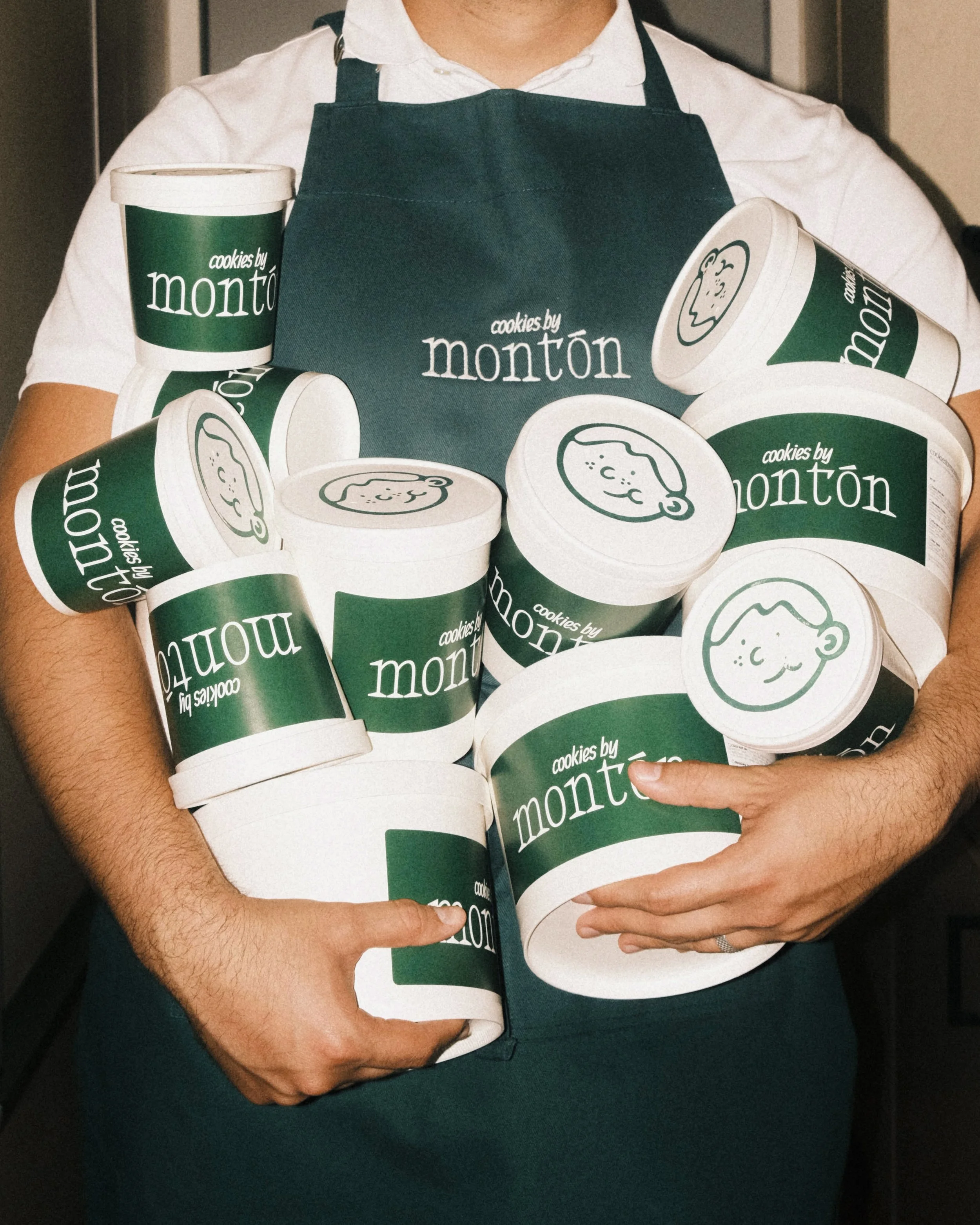 Person wearing a green apron holding multiple containers of cookies from Cookies by Montón.