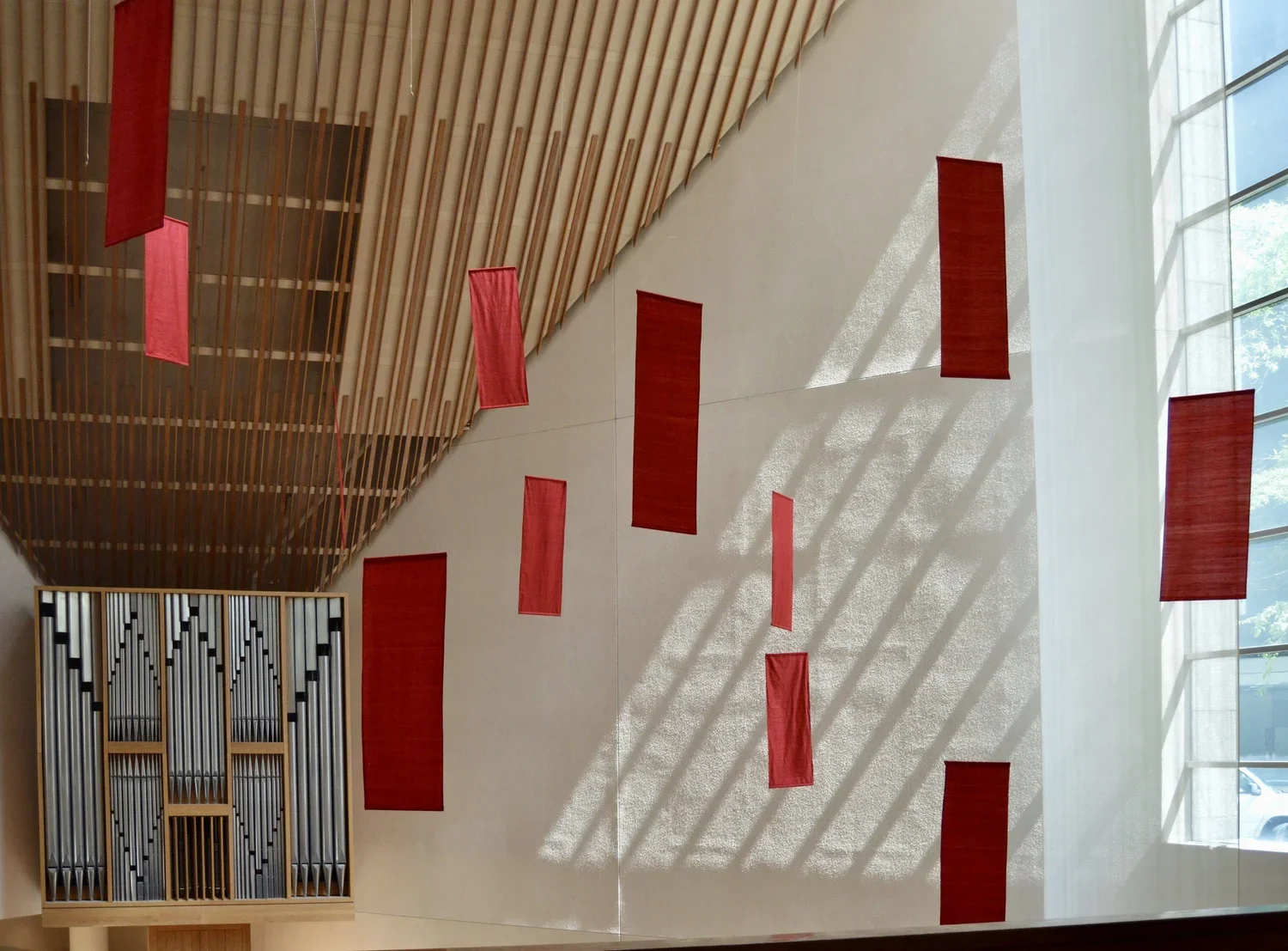 Interior of a modern building with a large white wall decorated with red fabric panels, a wooden ceiling with slats, and a large window with sunlight casting shadows.