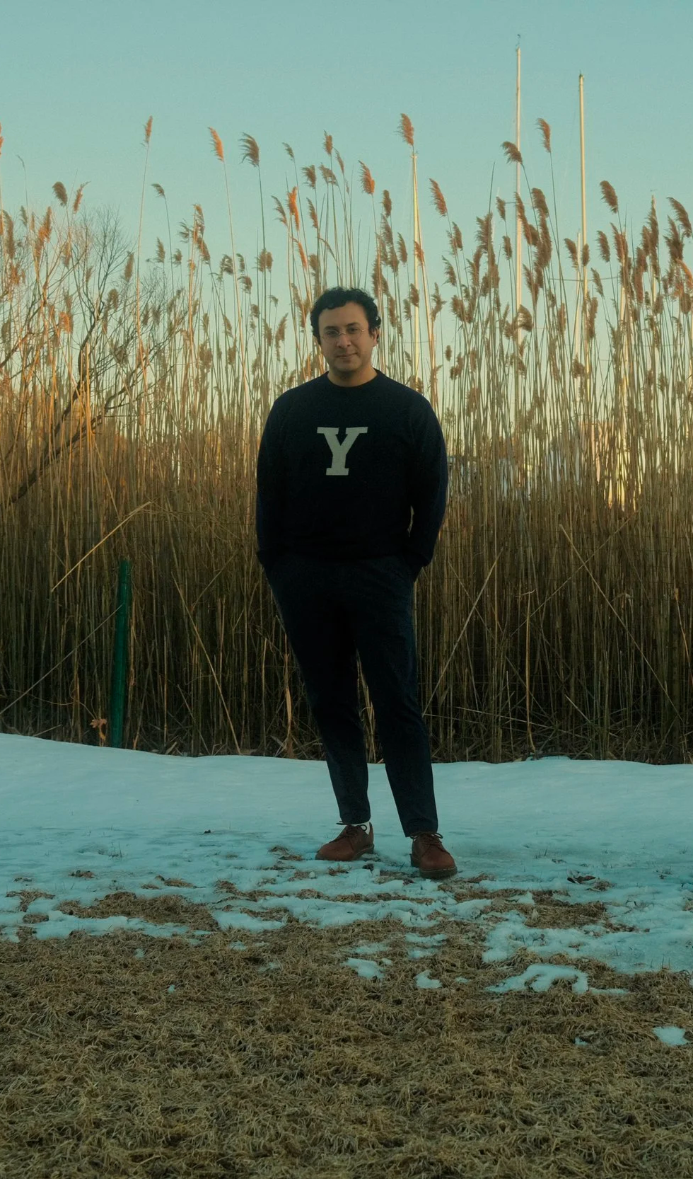 A man standing outdoors on a patch of snow-covered ground, with tall dried reeds or grass in the background, wearing a dark sweater with a large white letter 'Y' on the front.