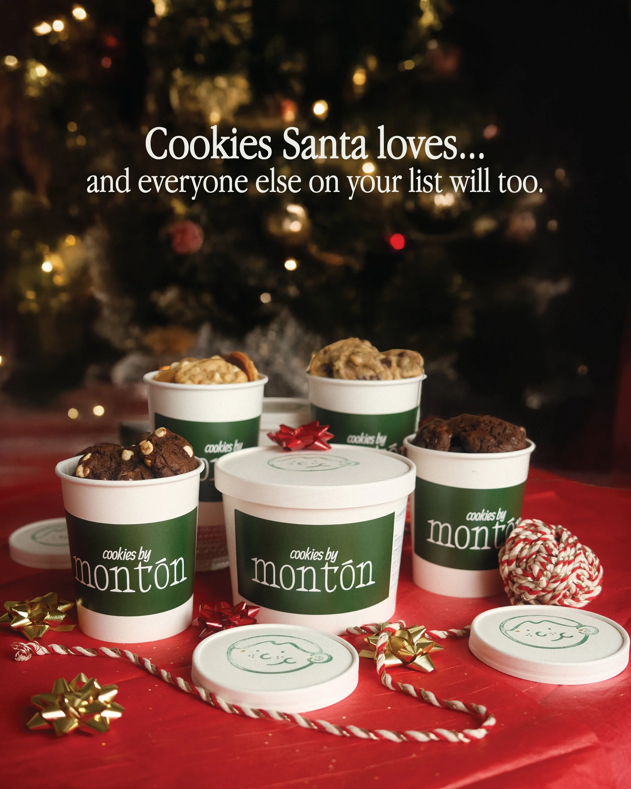 Assorted cookies in Montón-branded cups and containers sitting on a red surface, with Christmas decorations and a Christmas tree in the background.
