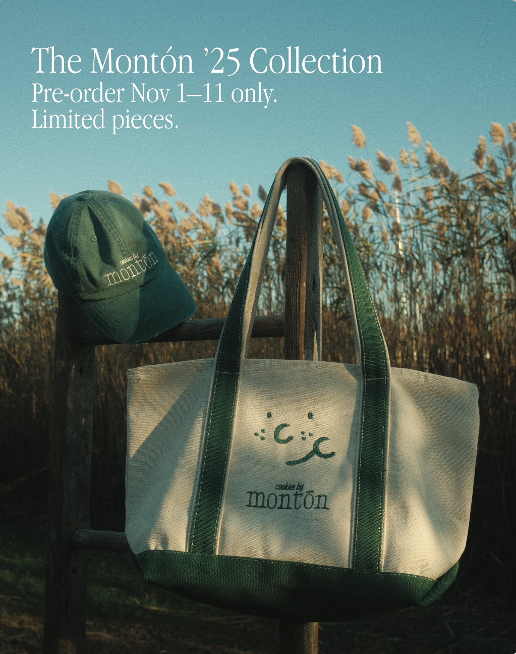 A green baseball cap and a beige tote bag with green straps hanging on a wooden post in a field of dry tall grass during the daytime. The bag has a cute face and the text 'cookies by montón' printed on it. The image promotes the Montón '25 Collection available for pre-order from November 1st to 11th.