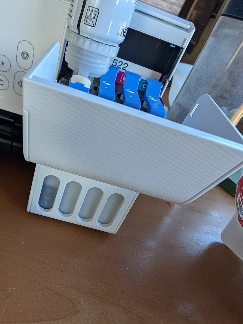 A white medication organizer with four pill compartments on a wooden surface, with colorful vials or ampoules stored inside, and a white device with buttons partially visible in the background.