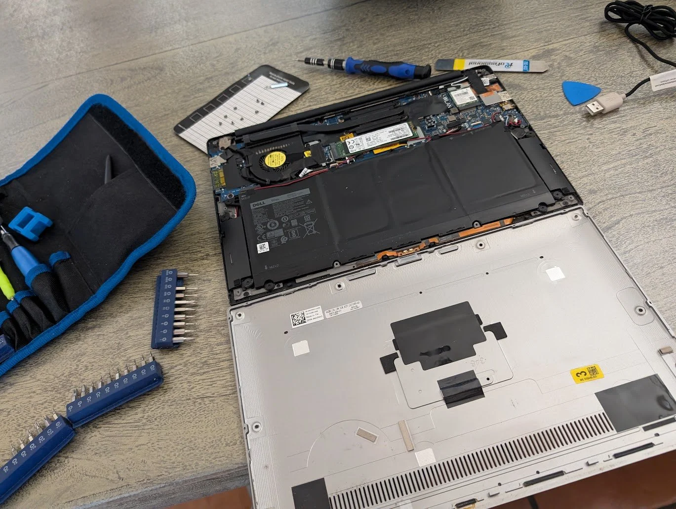 Open laptop with internal components exposed, repair tools nearby including screwdrivers, spudger, and a tool kit, on a wooden surface.