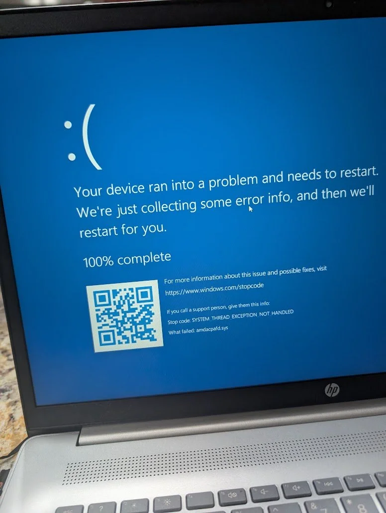 A laptop screen displaying a Windows blue screen error message with a sad face, indicating a system crash and restart needed, including a QR code and troubleshooting information.