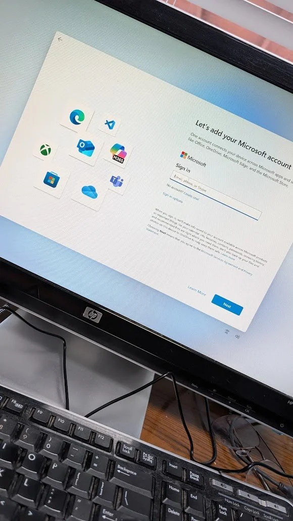 Computer monitor displaying a Microsoft account sign-in page with a selection of app icons, a keyboard, and a wooden desk.