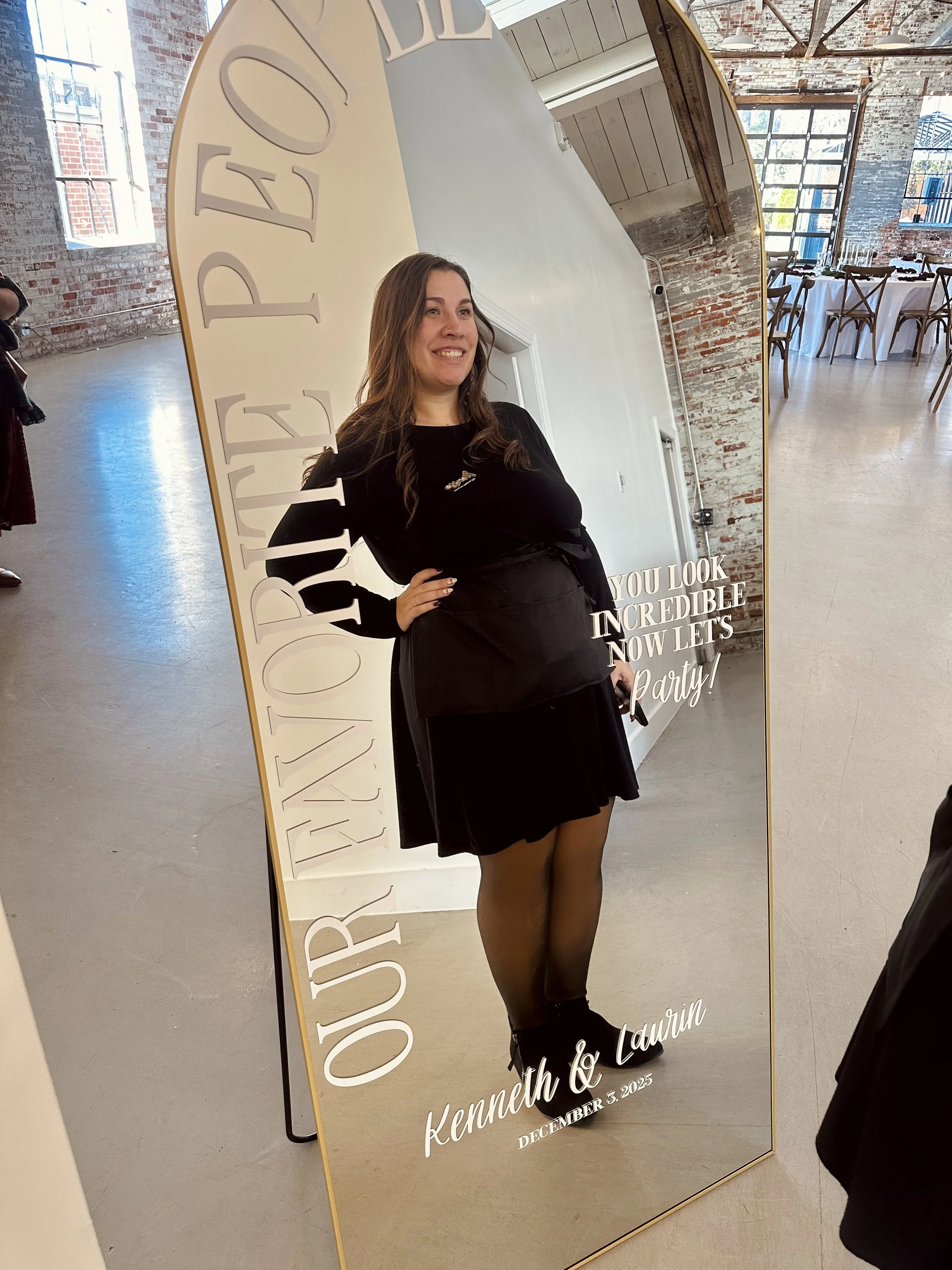 A woman looking at herself in a large decorative mirror at an event venue. The mirror has white text that reads, 'OUR JOURNEY FROM YOU LOOK INCRIDIBLE NOW LET'S Party! Kenneth & Laurin DECEMBER 5, 2025.' The woman is smiling, with long brown hair, wearing a black dress, sheer tights, and black shoes. The background shows a rustic, industrial interior with brick walls, large windows, and a dining area with tables and chairs.