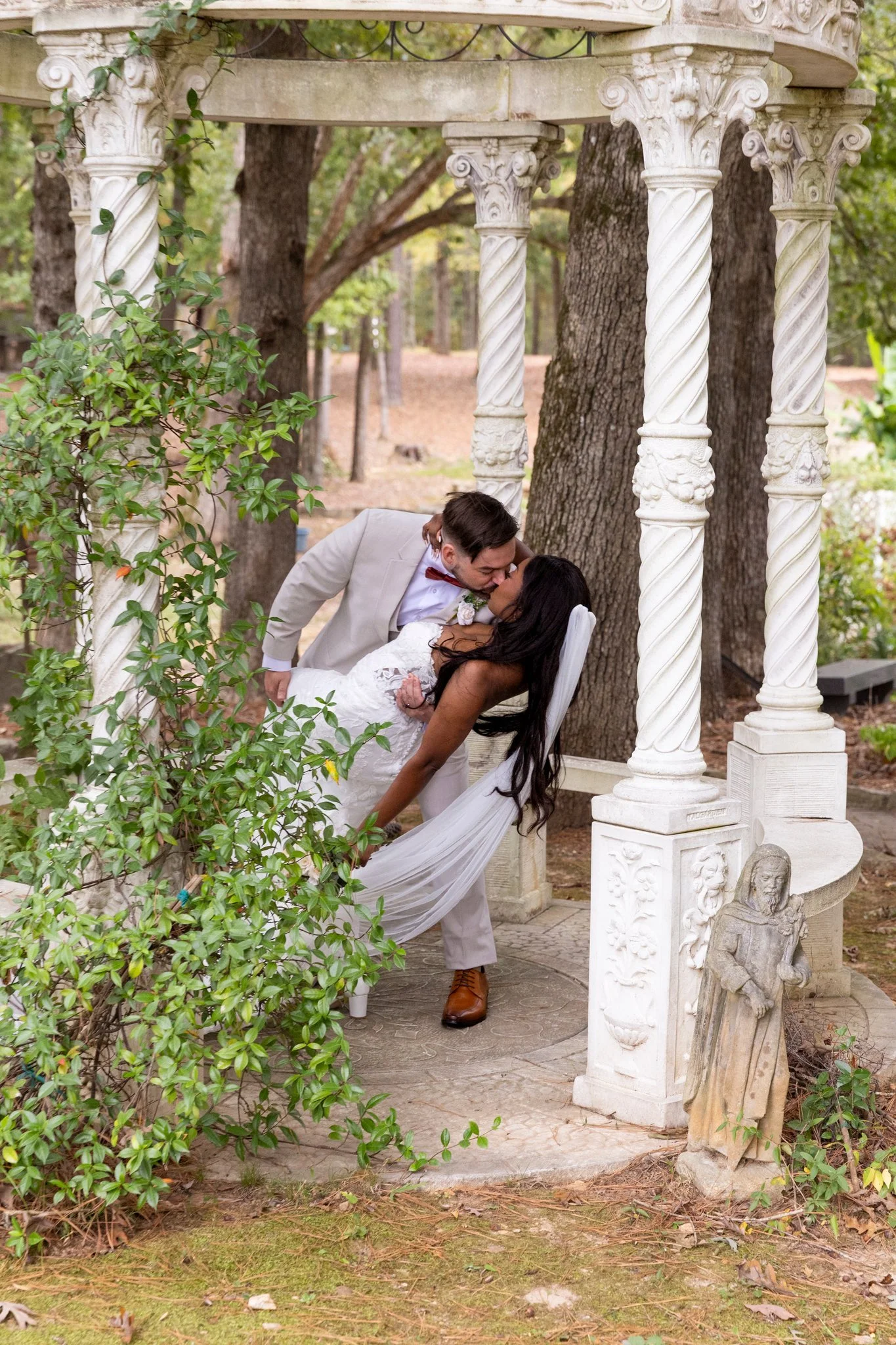 A newlywed couple kissing under a decorative white stone gazebo, surrounded by trees and greenery in a garden setting.