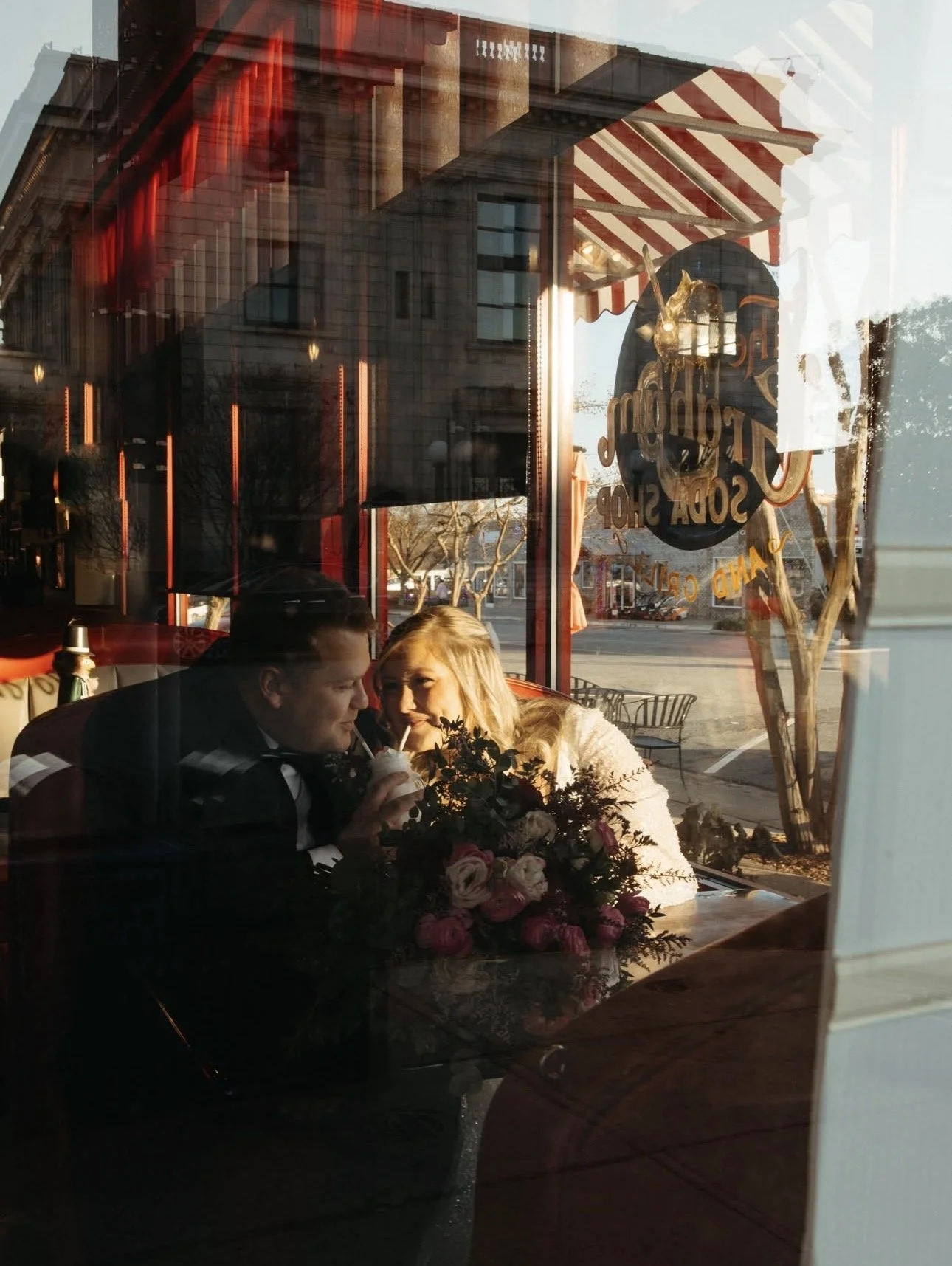 A couple sitting at a table inside a cafe, seen through the window, with the man in a tuxedo drinking from a straw and the woman in a white dress smiling, with a floral arrangement on the table, during sunset.