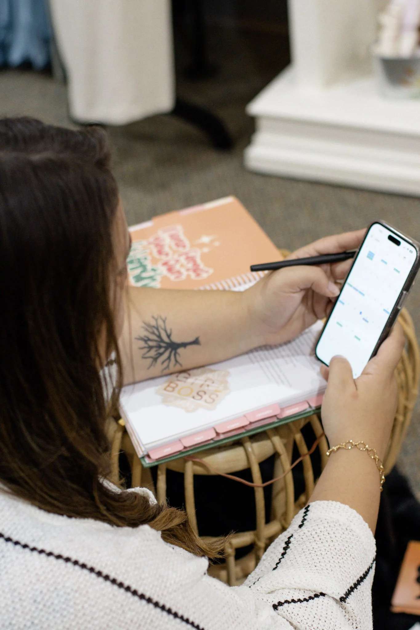A woman with a tattoo of a tree on her arm holding a stylus and looking at her smartphone with a stylus in hand, seated at a wicker table with a holiday-themed notebook.