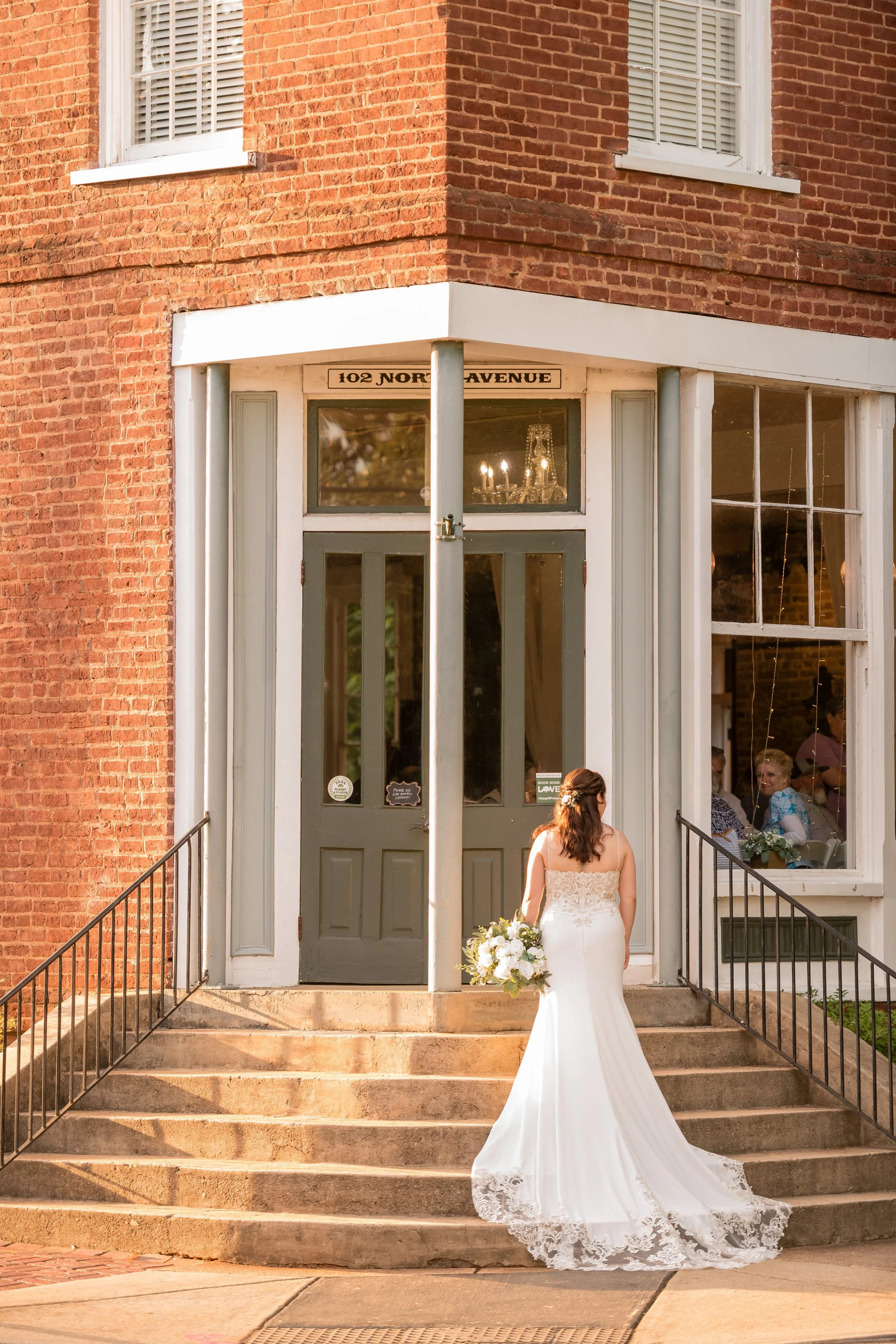 A bride in a white wedding dress holding a bouquet stands on the steps of a brick building with large windows and dark gray doors, with the address 102 North Avenue visible above the door, as people sit inside through the windows.