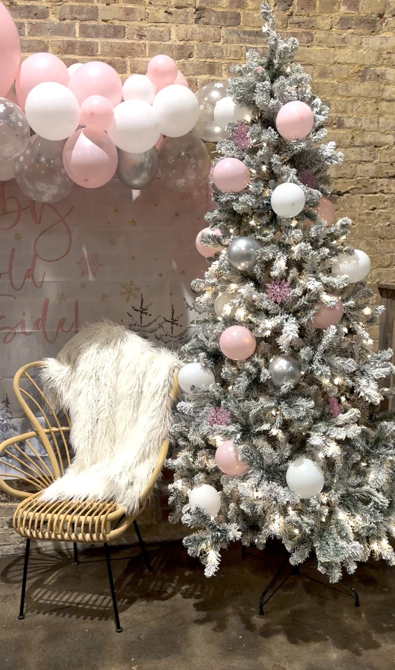A decorated Christmas tree with pink, white, and silver ornaments, next to a rattan chair with a white furry blanket, against a brick wall and a balloon garland in pastel pink, white, and silver.
