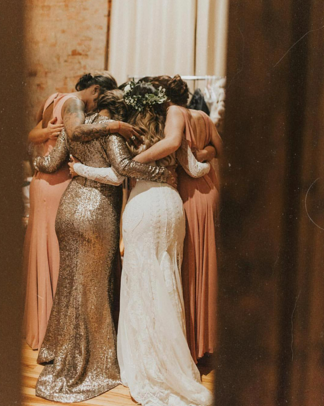 Five women dressed in elegant dresses share a group hug indoors.