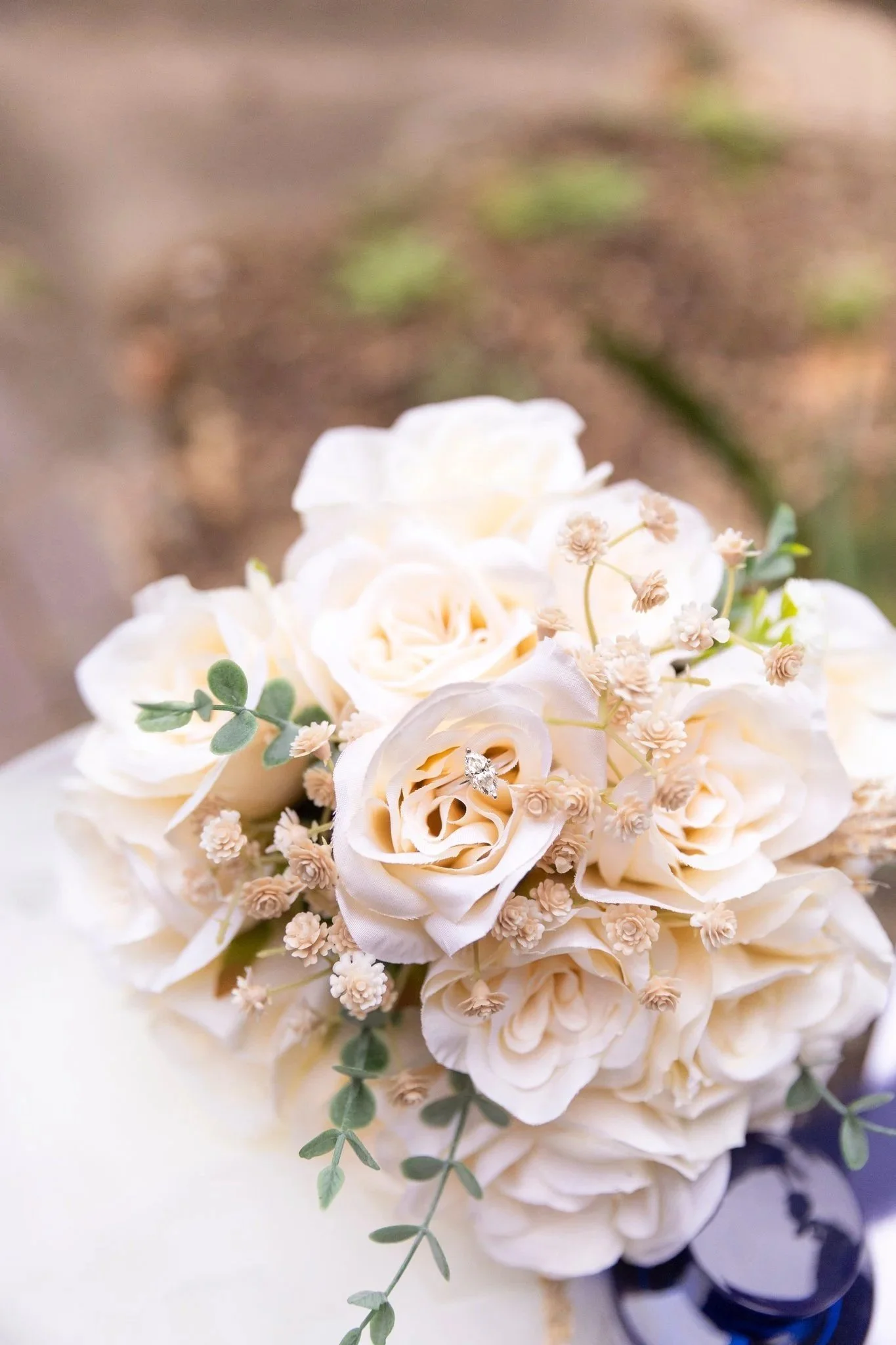 A bouquet of white roses with small white flowers and green leaves, with a diamond ring placed inside one of the roses.