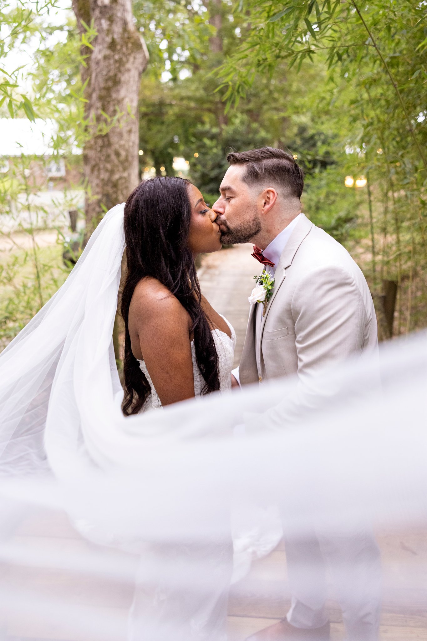 A newlywed couple sharing a kiss outdoors on a wooden pathway surrounded by trees, the bride wearing a white wedding gown and veil, and the groom in a light-colored suit and red bow tie.