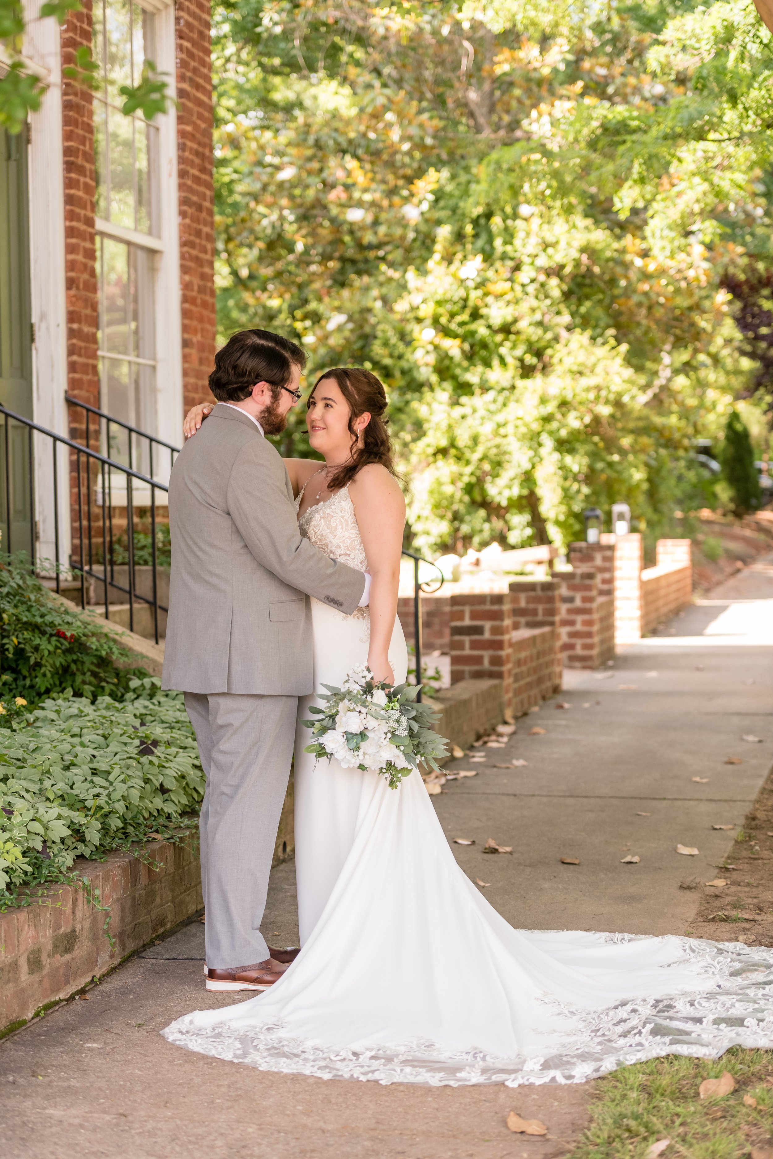 A newlywed couple standing on a sidewalk outside a brick building with greenery, embracing and looking at each other, with the bride holding a bouquet of white flowers and greenery.