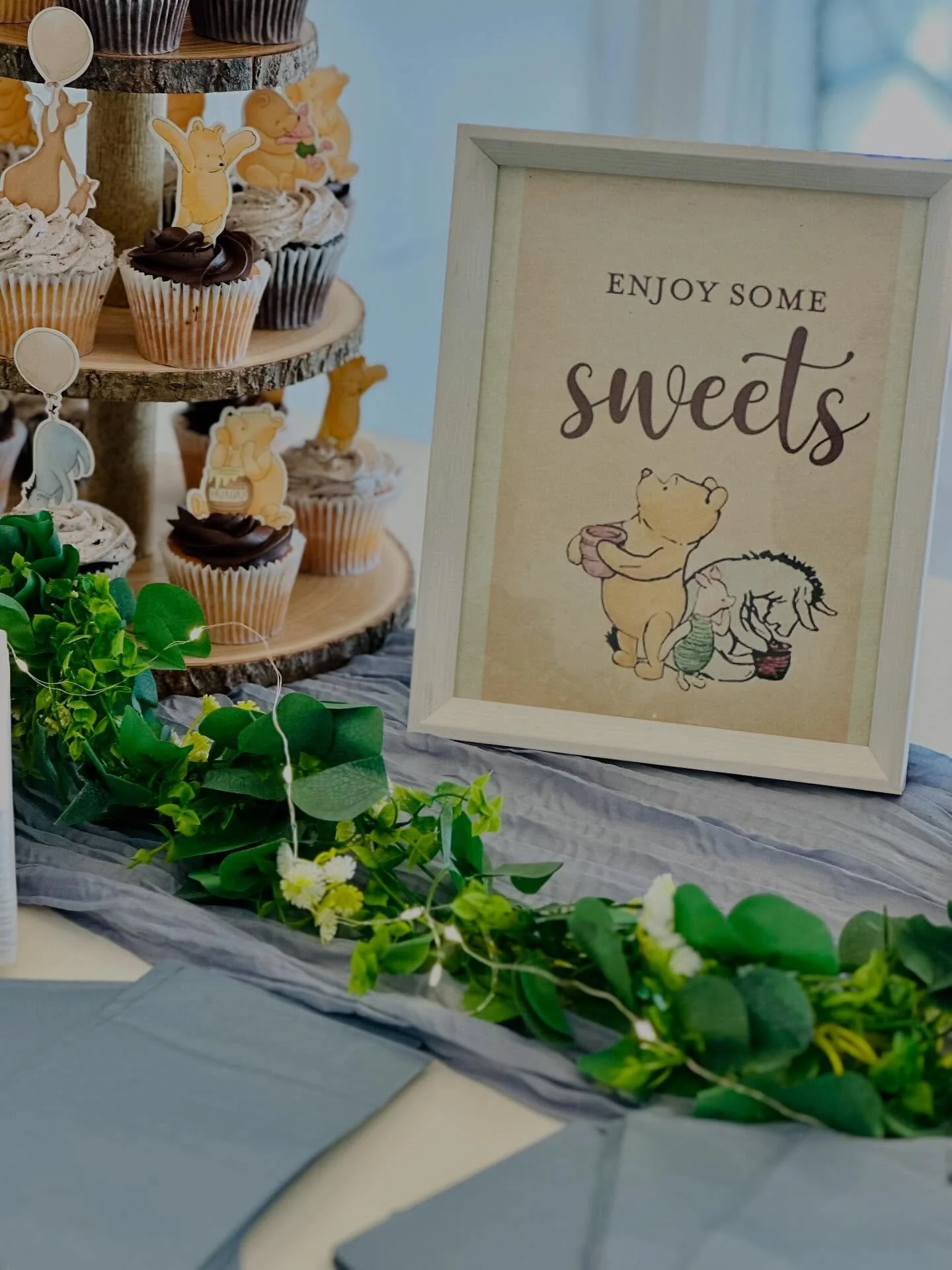 A framed sign reading 'Enjoy some sweets' with a Winnie the Pooh illustration, set on a table decorated with green leaves and a cupcake display with various cute animal toppers.