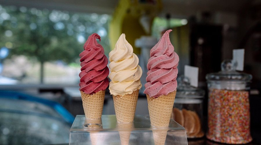 Three soft serve ice cream cones with twisted scoops, from left to right: red, white, and pink, displayed on a clear stand in an ice cream shop.