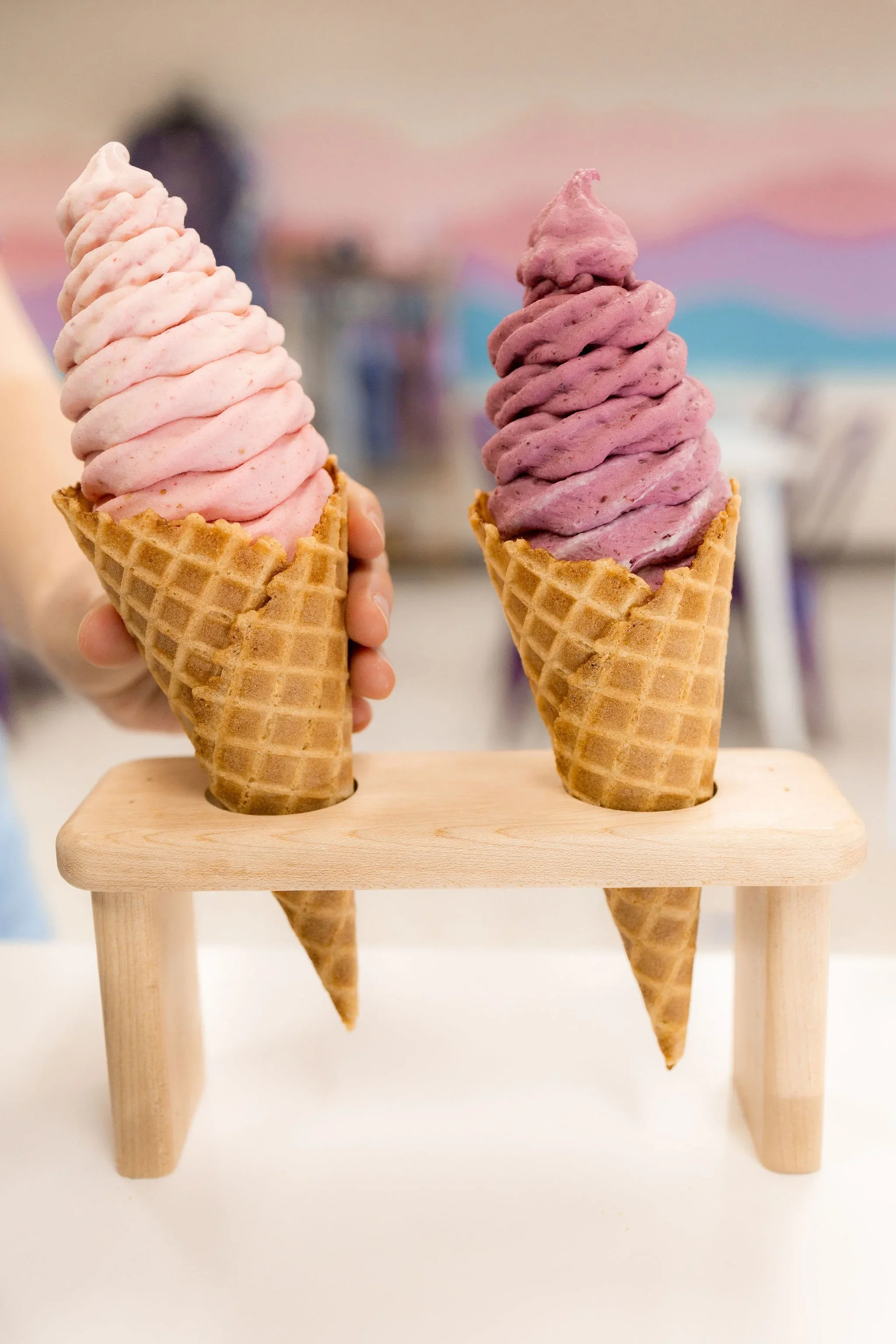 Two soft serve real fruit frozen yogurt ice cream cones, one pink and one purple, held in front of a colorful background, on a small wooden stand.